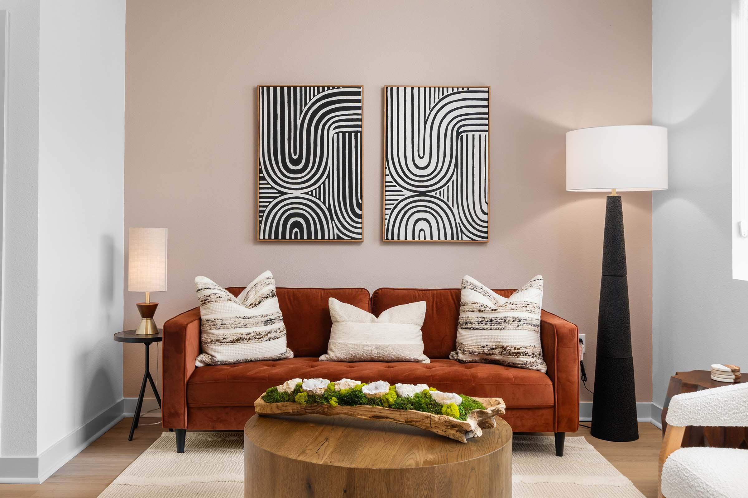 A cozy living room featuring a rust-colored sofa adorned with patterned pillows. Two abstract black and white artworks hang on the wall above the sofa. A modern floor lamp stands beside the couch, and a wooden coffee table with decorative items sits in front of it, all set against a light, neutral wall.