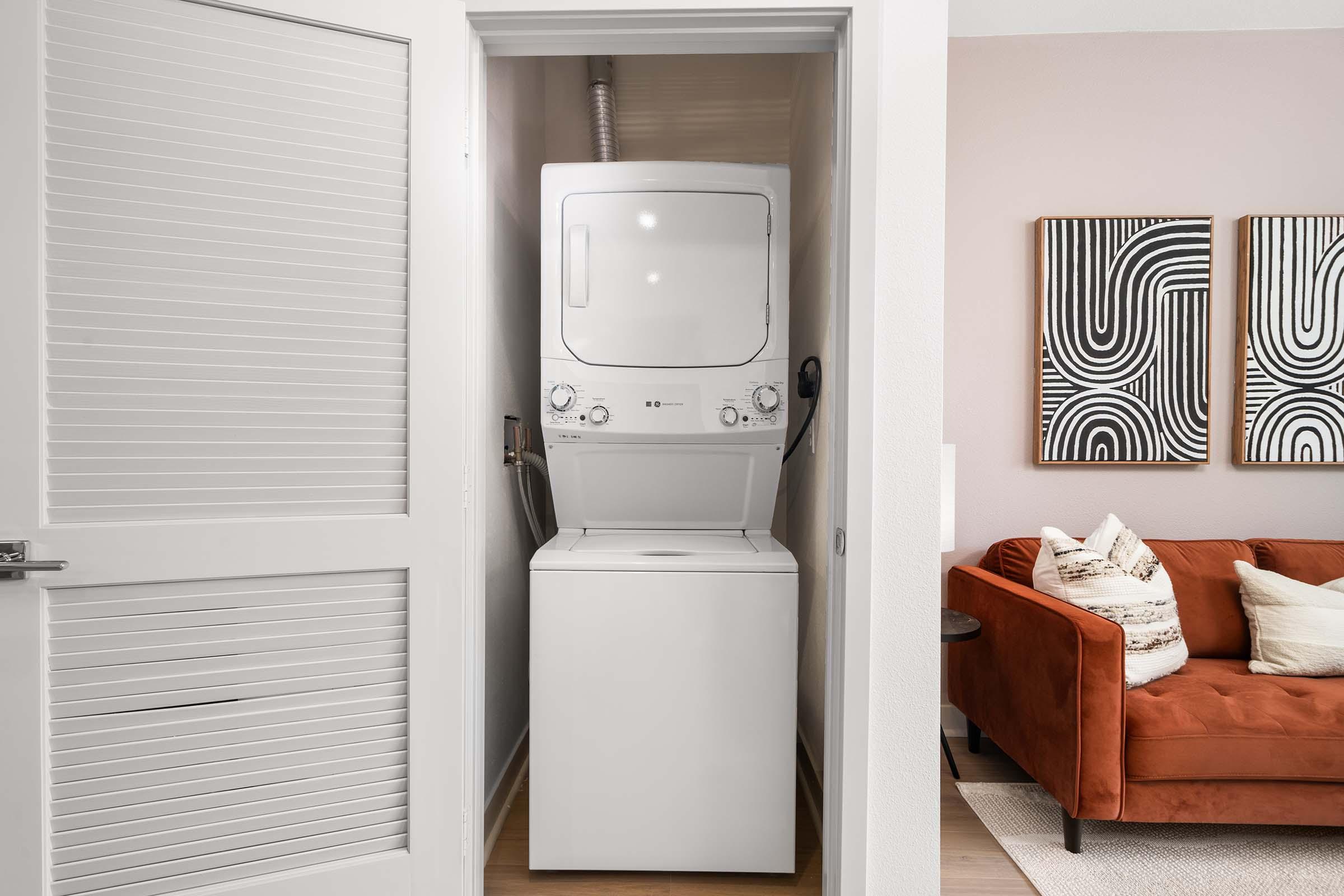 Stacked washer and dryer located in a small laundry area enclosed by a door; a cozy living room is visible in the background, featuring a modern orange couch and abstract wall art.
