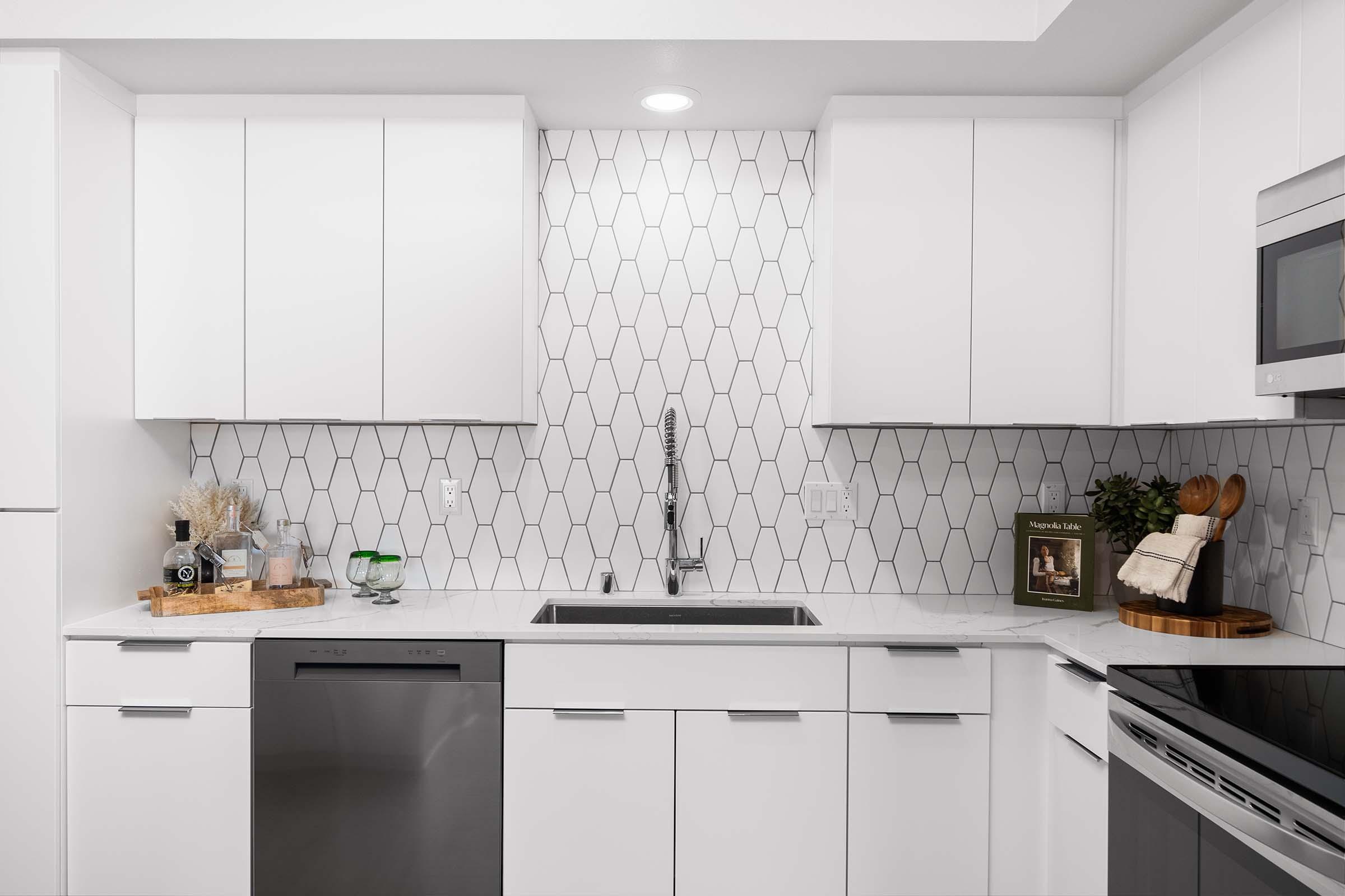 Modern kitchen with white cabinetry and stainless steel appliances. The backsplash features a hexagonal tile pattern, and the countertop is light-colored. A sink with a sleek faucet is centrally located, accompanied by decorative items like a wooden cutting board and plants.