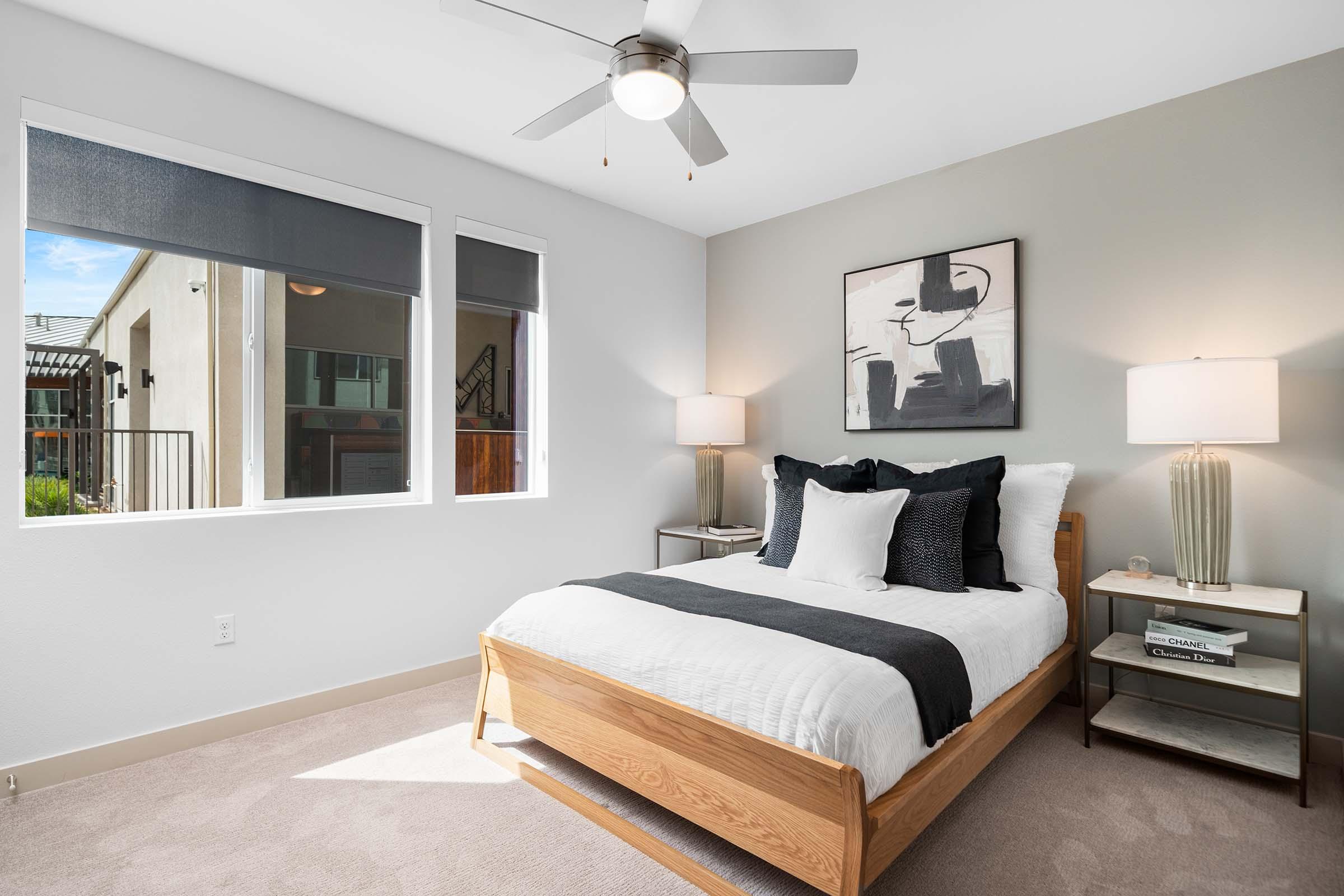 A cozy bedroom featuring a modern design with a wooden bed frame, white bedding, and black accent pillows. The room includes two bedside lamps, a minimalist piece of wall art, and large windows allowing natural light in. A ceiling fan is visible, enhancing the airy atmosphere.