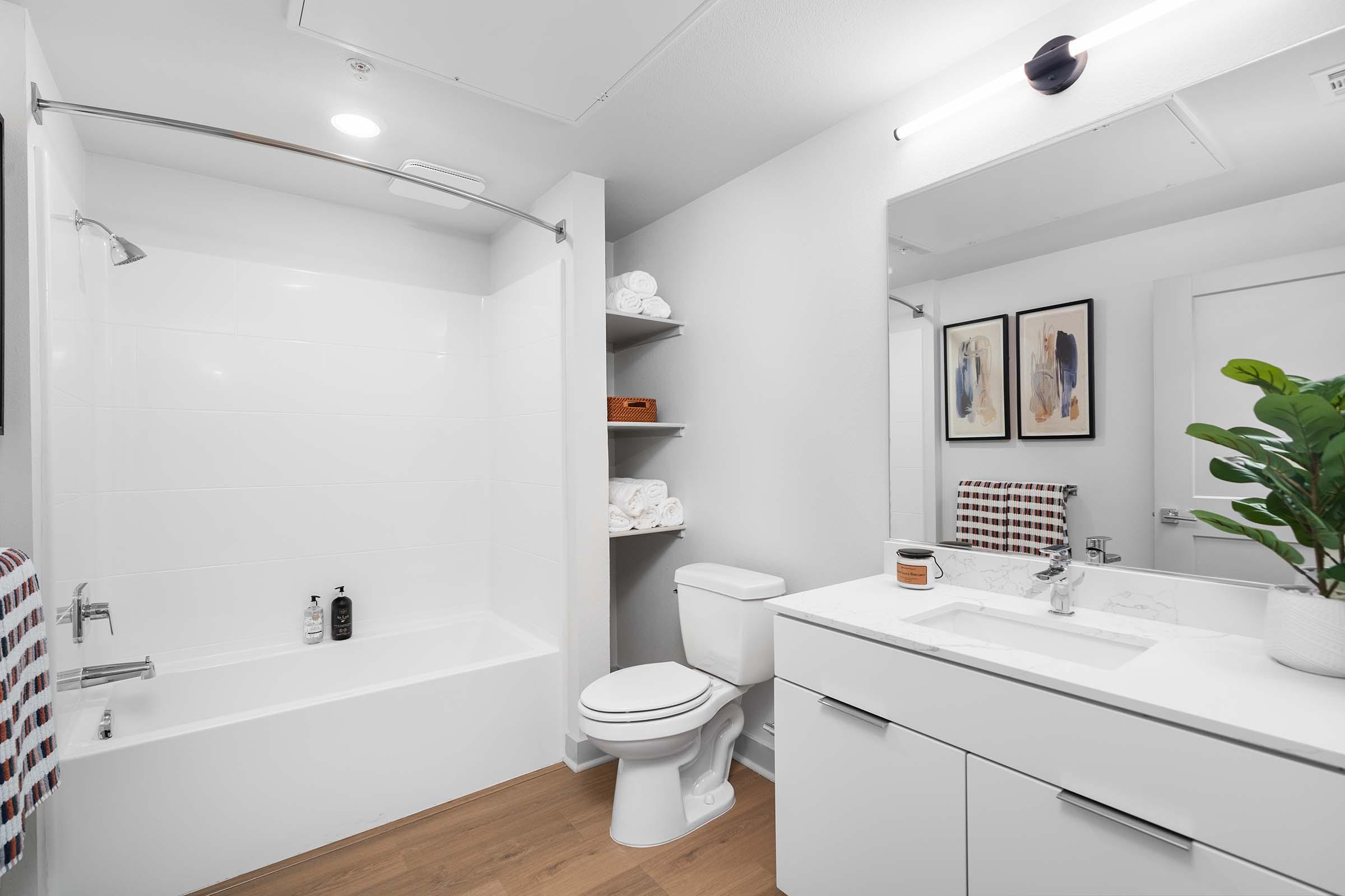 A modern bathroom featuring a white shower/tub combination, a toilet, and a sleek vanity with a sink. The vanity is equipped with storage and a mirror. A small plant adds a touch of greenery, while neatly stacked towels are visible in the background, accompanied by framed artwork on the wall. The flooring is light wood.