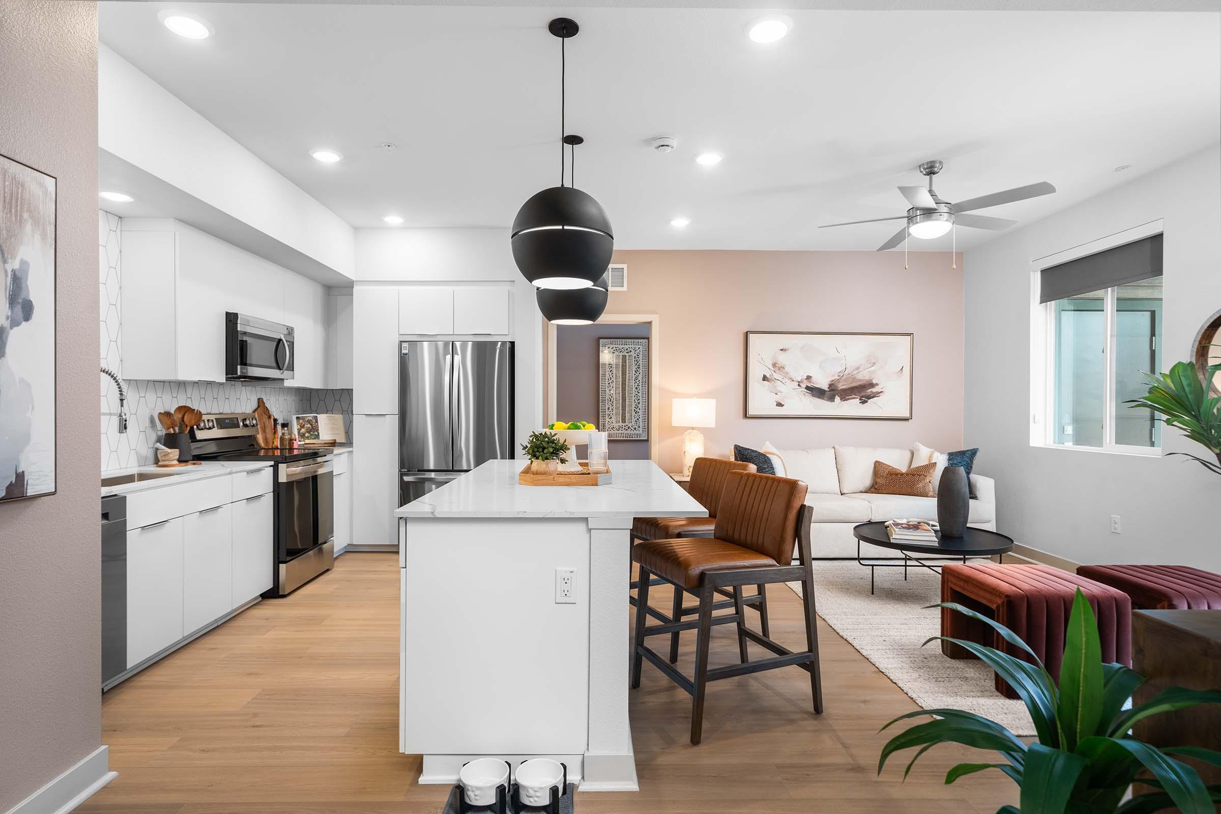 Modern kitchen and living area featuring a marble-top island with seating, stainless steel appliances, and a decorative wall painting. The living space includes a sofa, accent chairs, and a plant, with soft lighting and a ceiling fan. The decor combines neutral tones with pops of color for a cozy ambiance.