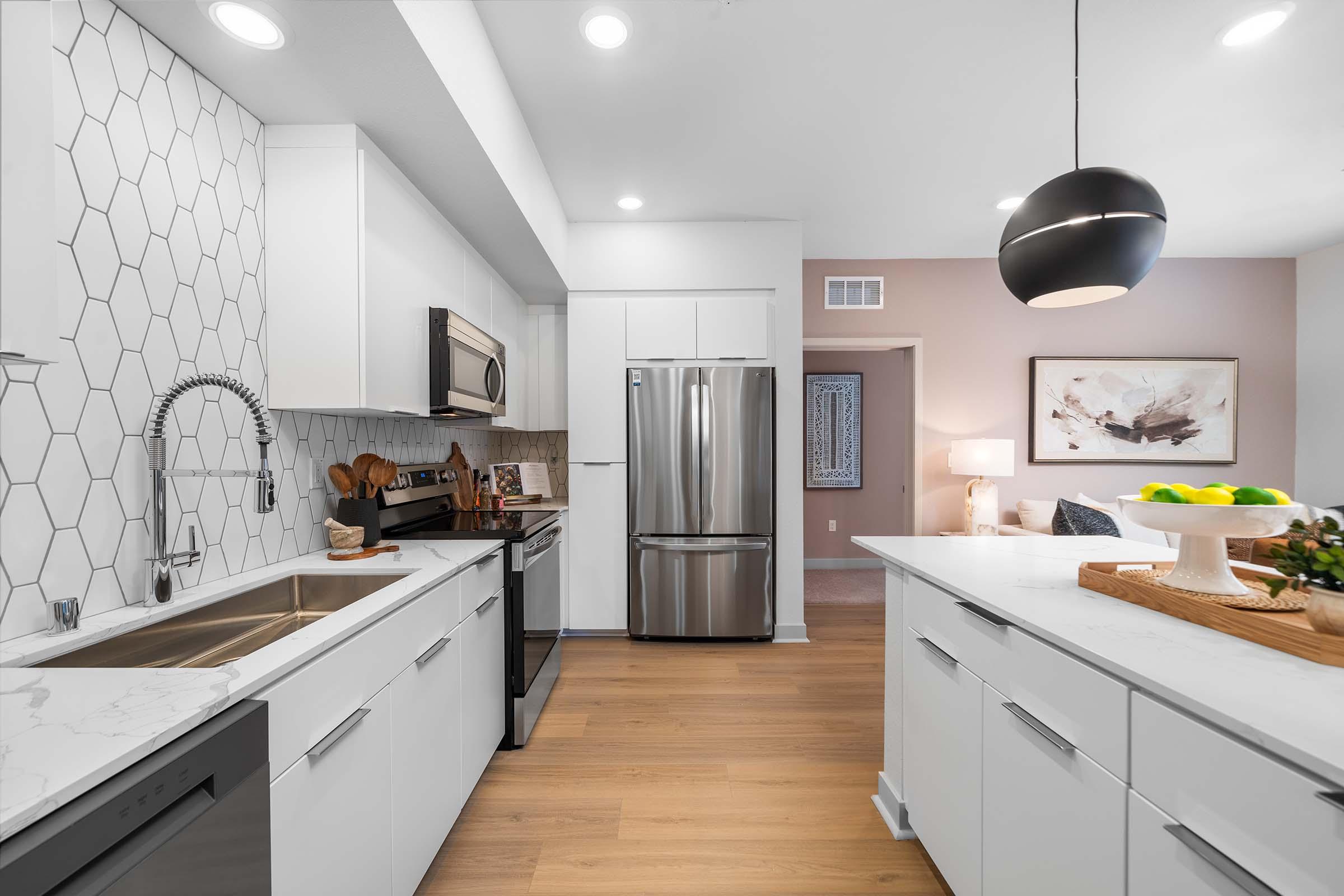 Modern kitchen featuring white cabinets, a geometric tile backsplash, stainless steel appliances, and a large island with a fruit bowl. The space is well-lit with recessed lighting and includes wooden accents, creating a warm and inviting atmosphere.