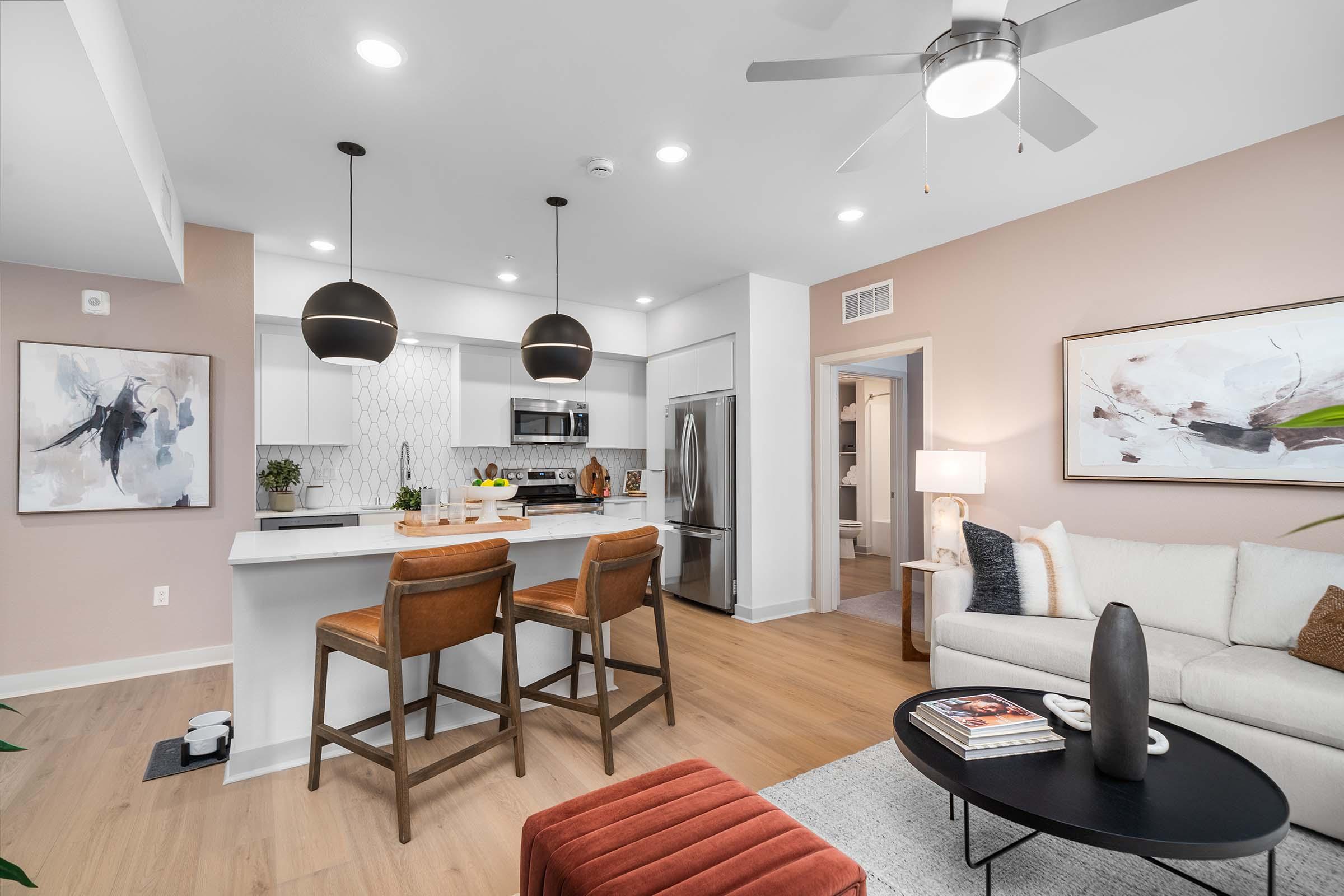Modern apartment interior featuring a kitchen with white cabinets, stainless steel appliances, and a central island with two wooden bar stools. A cozy living area includes a cream-colored sofa, an accent ottoman, and artwork on the walls. Soft lighting and neutral tones create a welcoming atmosphere.