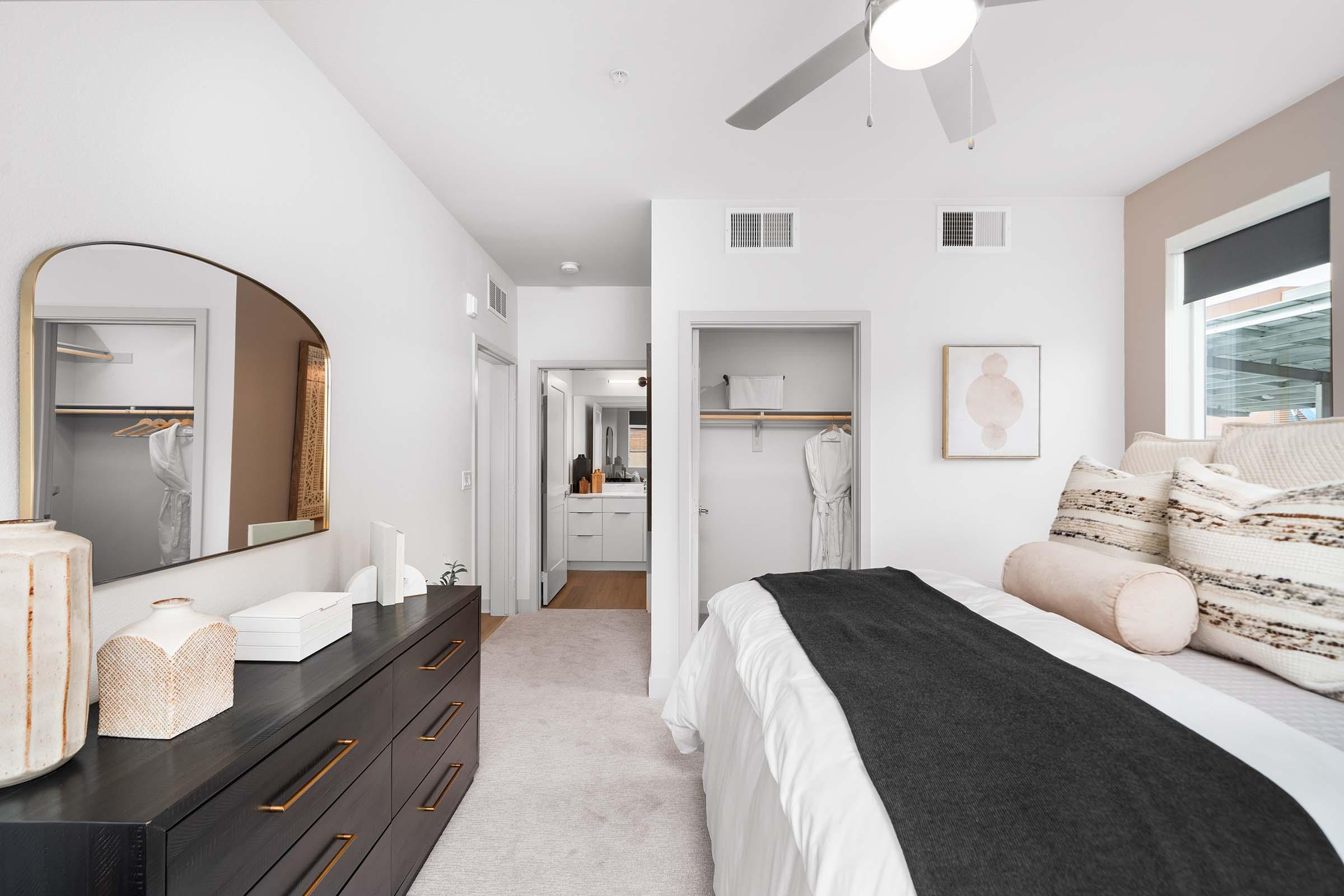 A modern bedroom featuring a large bed with a dark throw blanket, decorative pillows, and a stylish headboard. There's a wooden dresser with decorative items, a large mirror on the wall, and an open closet. In the background, a bathroom area is visible with bright, clean lines and neutral colors. Natural light illuminates the space.