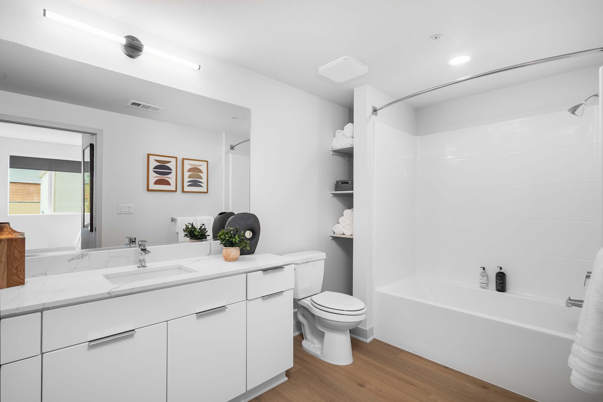 Modern bathroom featuring a clean and bright design. It includes a white bathtub with a shower, a double sink vanity with minimalistic fixtures, and neatly arranged towels on a shelf. Wall art adds a touch of elegance, and there is plenty of natural light coming from a window. Wood flooring completes the contemporary look.