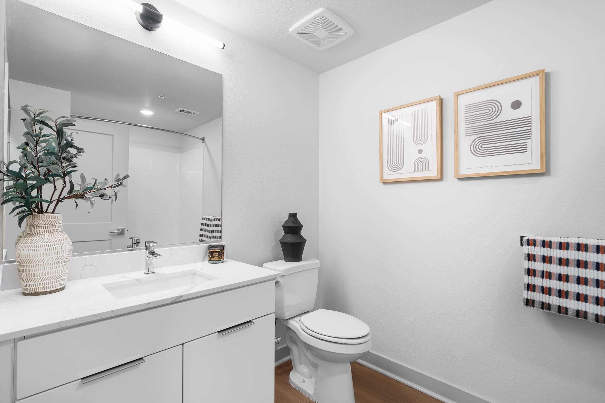 A modern bathroom featuring a white sink and vanity with a marble countertop, a toilet, and a shower area. Two framed abstract art pieces hang on the wall. A decorative vase with greenery sits on the countertop, and a striped towel hangs nearby, complementing the minimalist design.