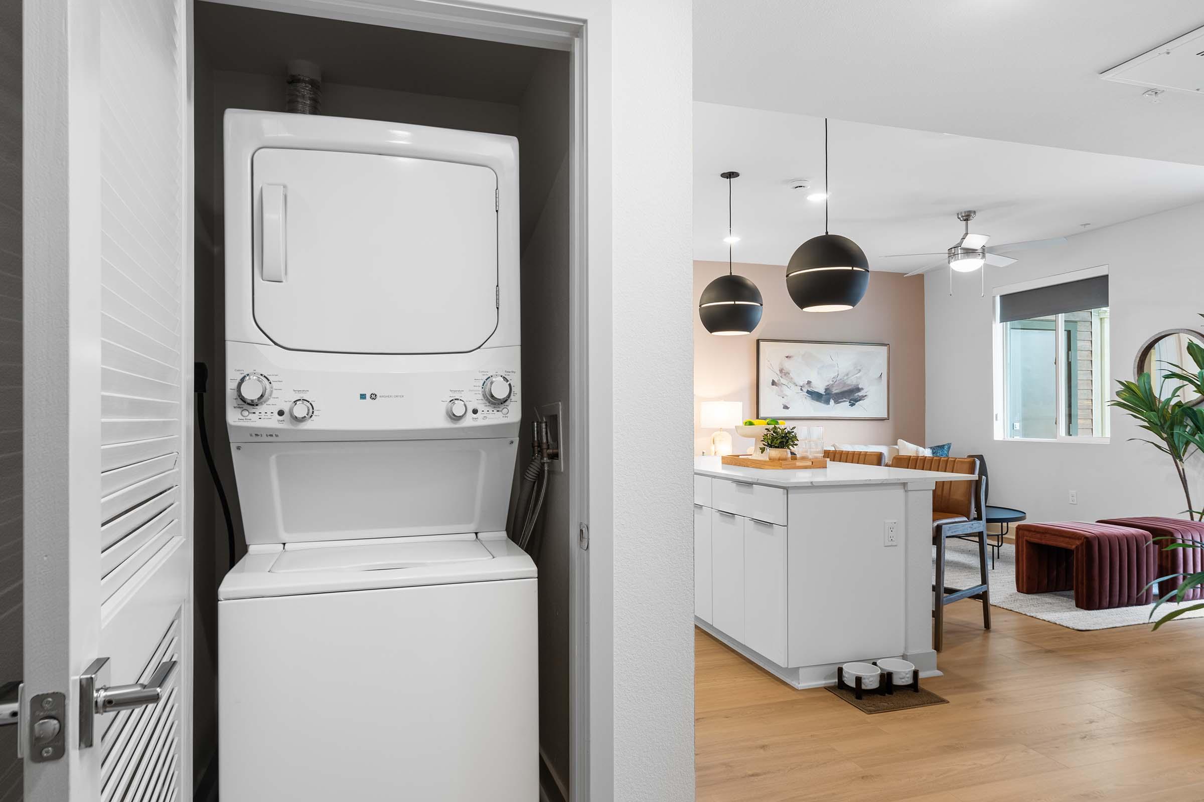 A stacked washer and dryer unit is positioned in a small laundry area. Beyond, a modern living space is visible, featuring a kitchen island with seating, pendant lights, and a cozy seating area with a coffee table. Natural light enters through a window, creating a bright and inviting atmosphere.
