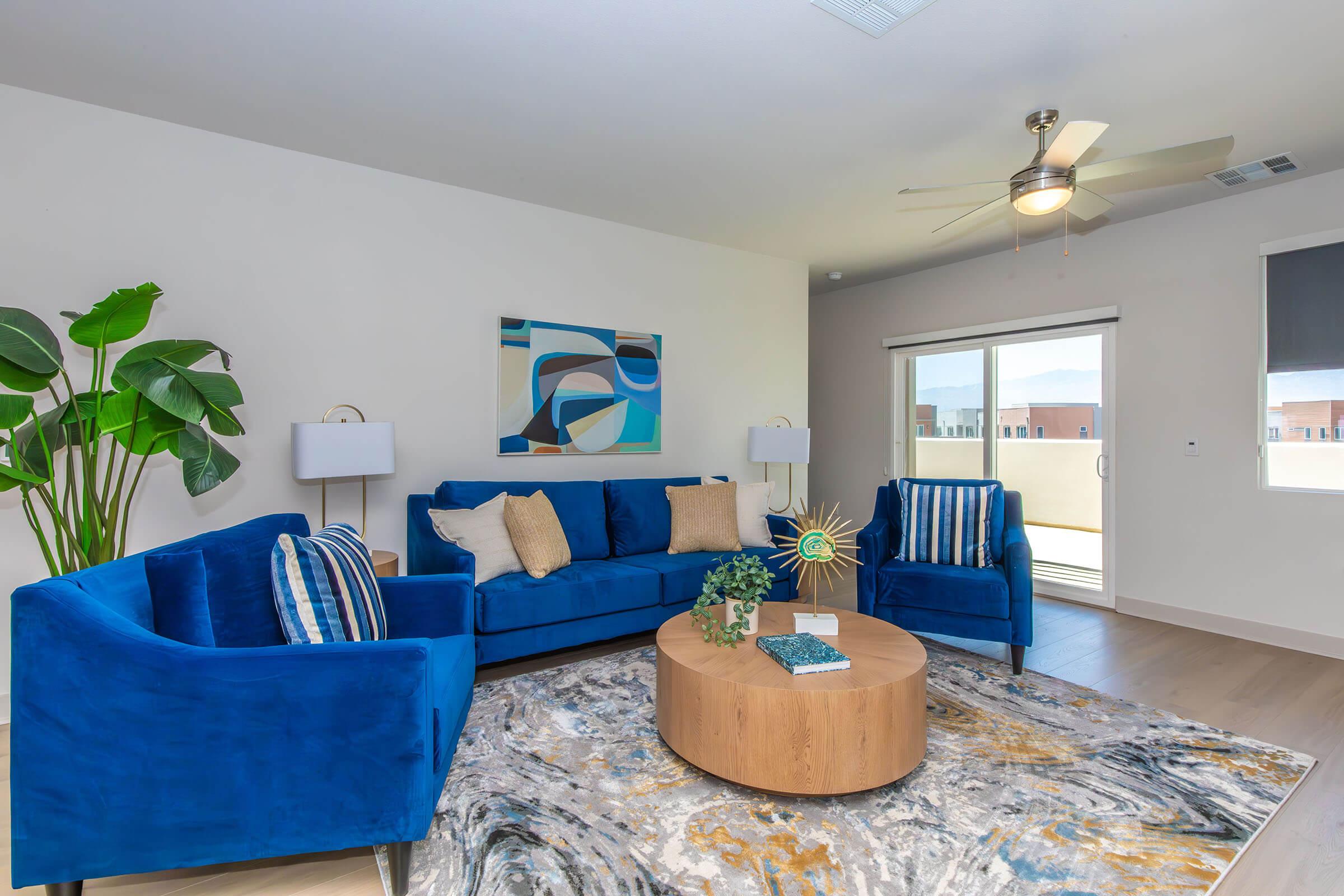 A modern living room featuring a blue sofa set with pillows, a round wooden coffee table, and a colorful area rug. There is a ceiling fan and large sliding glass doors leading to a balcony. Decorative plants and artwork adorn the space, creating a stylish and inviting atmosphere.