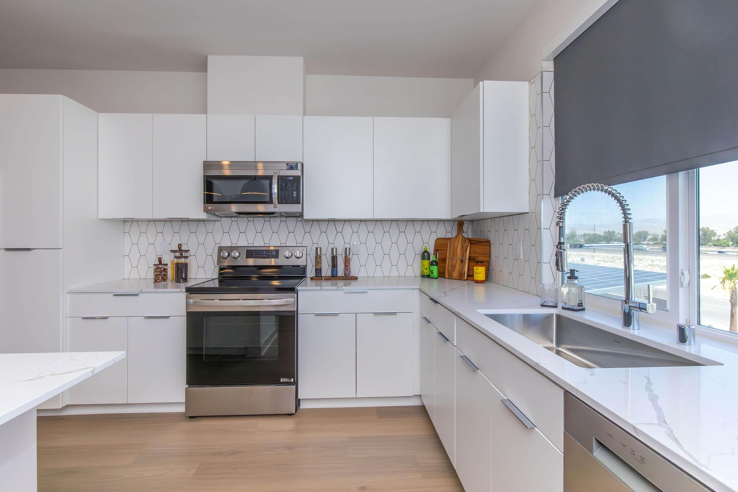 Modern kitchen featuring sleek white cabinetry, stainless steel appliances including a microwave and oven, a minimalist backsplash with geometric tile design, and a spacious countertop with a double sink. Natural light streams in through a large window with a gray roller shade.