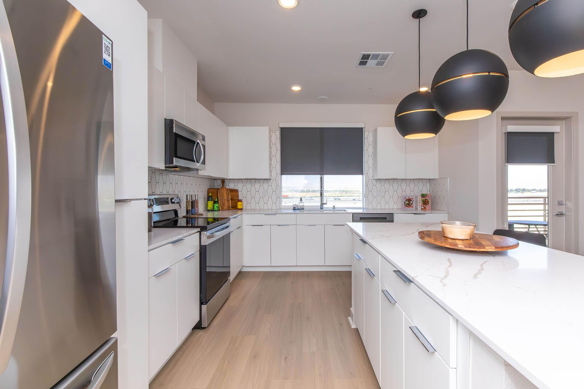 Modern kitchen featuring stainless steel appliances, a large island with a wooden serving tray, elegant pendant lights, sleek white cabinetry, and a patterned backsplash. Sunlight streams through large windows, providing a bright and airy atmosphere. The overall design is contemporary and functional.