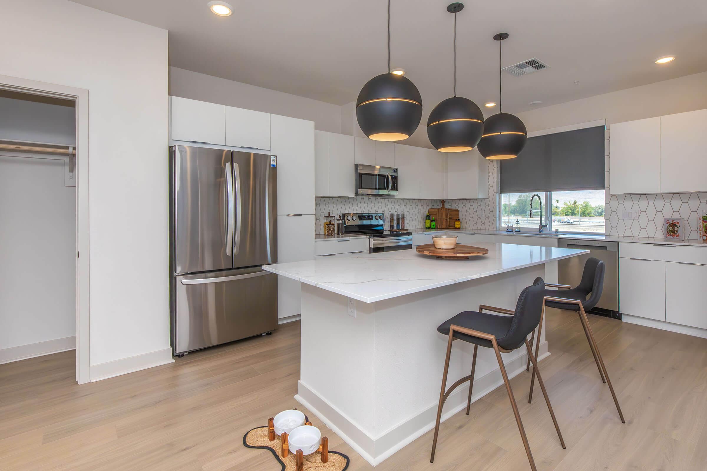 A modern kitchen featuring a white island with dark stools, stainless steel appliances, and geometric tile backsplash. Pendant lights hang above the island, and there's a window with a view. The floor is light wood, and there are decorative items on the countertop.