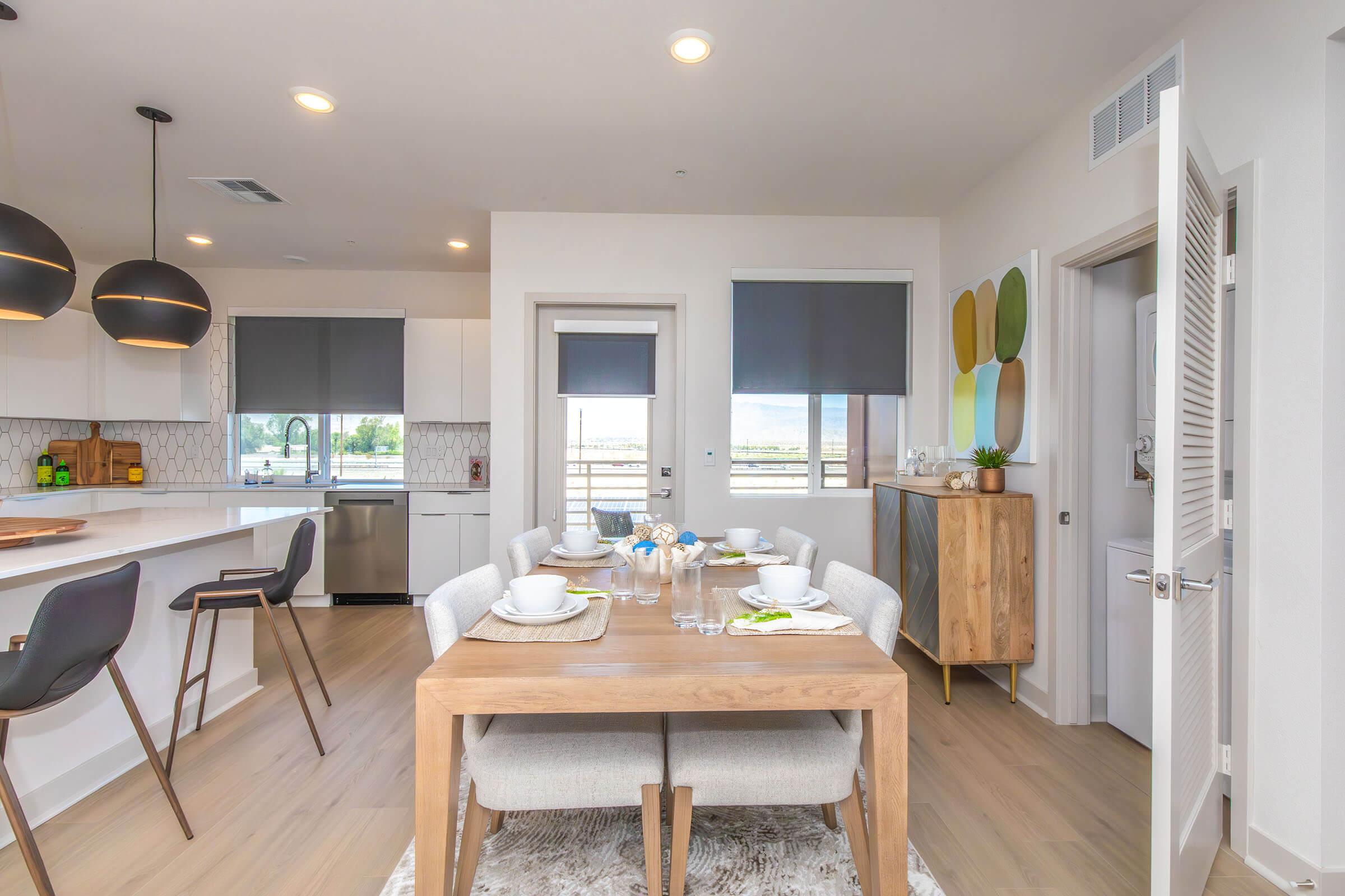 A modern dining area with a wooden table set for a meal, featuring white dishes and blue flowers. The kitchen in the background has white cabinets and a spacious layout. Large windows provide natural light, and decorative elements include artwork on the wall and stylish light fixtures above the table.