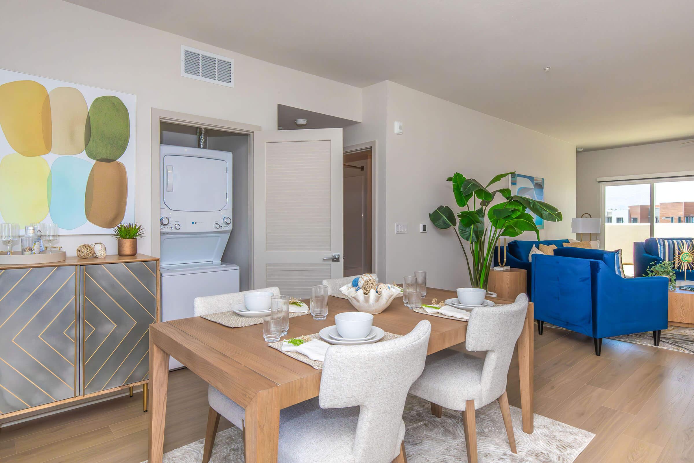 A modern dining area with a wooden table set for four, featuring plates and glasses. In the background, there is a laundry space with a stacked washer and dryer. Stylish plants and blue chairs add color to the room, which has neutral walls and a decorative wall art piece with abstract shapes.