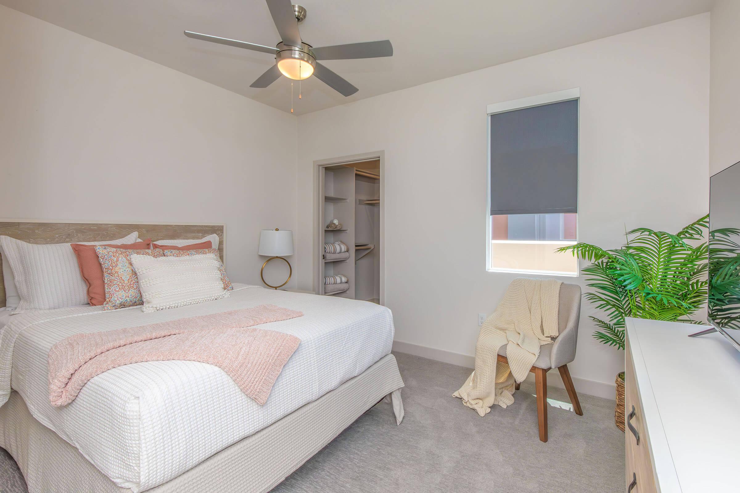 A cozy bedroom featuring a comfortable bed with decorative pillows and a soft throw blanket. A ceiling fan hangs above, and there is a lamp beside the bed. A plant adds a touch of greenery, while a small chair and a media console with a TV complete the room design. A closet is visible in the background.