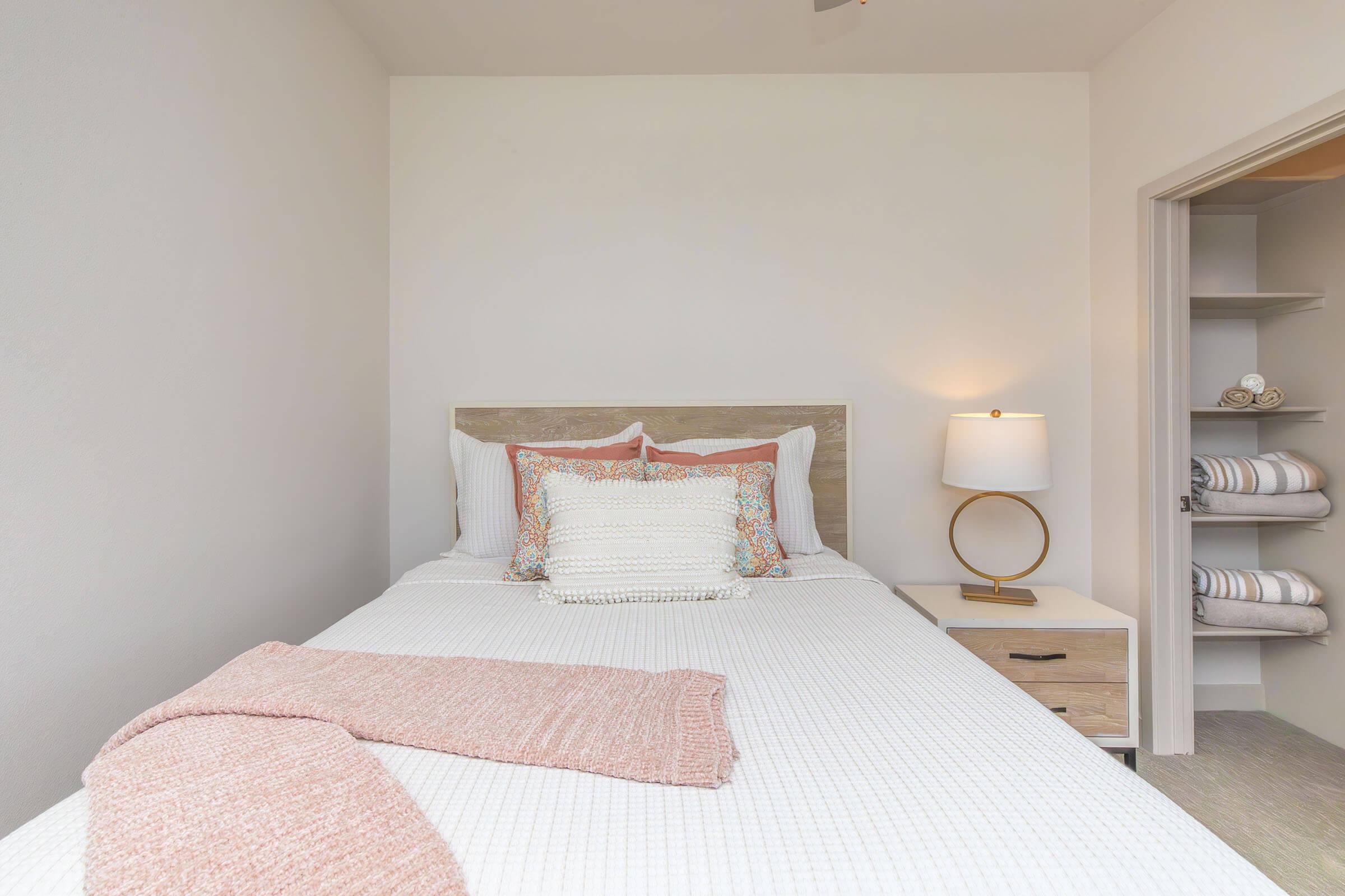A cozy bedroom featuring a neatly made bed with decorative pillows and a textured blanket. A bedside table with a lamp sits next to the bed, and an open closet with neatly arranged linens is visible in the background. The walls are painted in soft, neutral tones.
