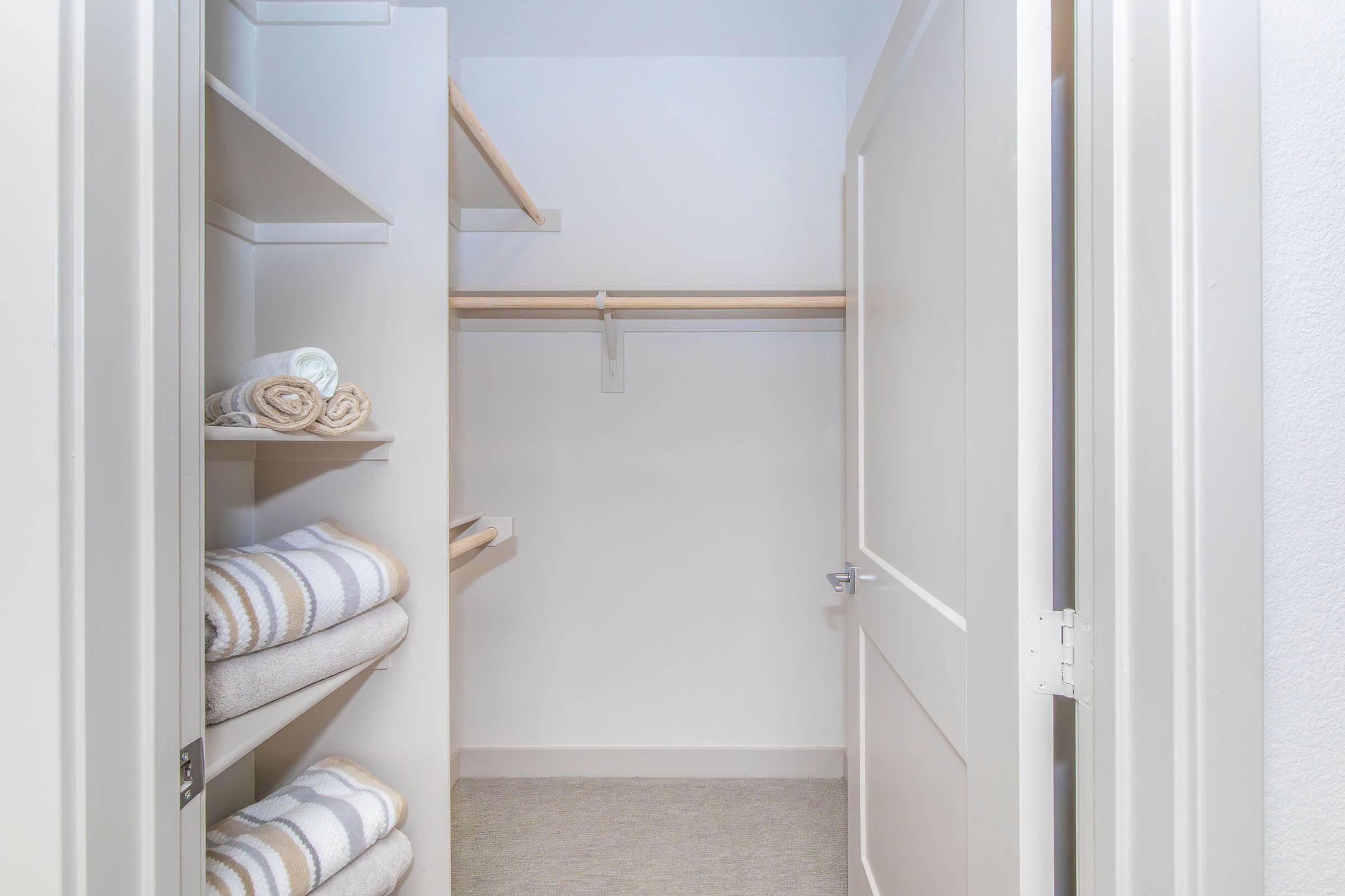 A clean, organized closet interior featuring light-colored walls. One side has shelves with neatly folded towels, while the opposite side features a hanging rod. The floor is covered with a soft carpet, and there is a door ajar, inviting natural light into the space.