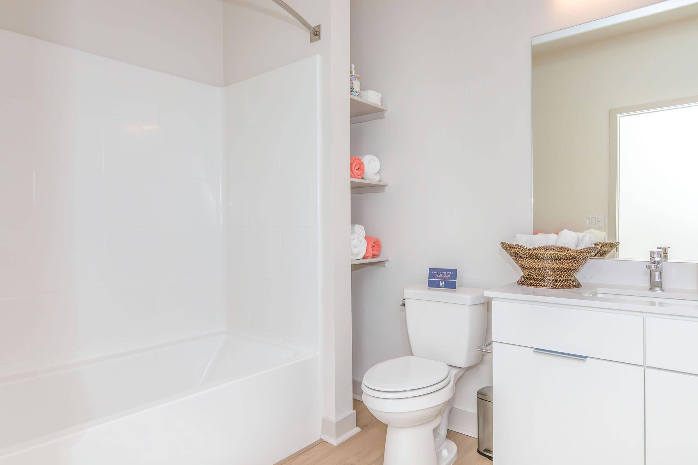 A clean and modern bathroom featuring a white bathtub, a toilet, and a vanity with a sink. The shelves hold neatly rolled towels, and there is a woven basket on the counter. Natural light enters through a nearby mirror, enhancing the bright and spacious feel of the room.