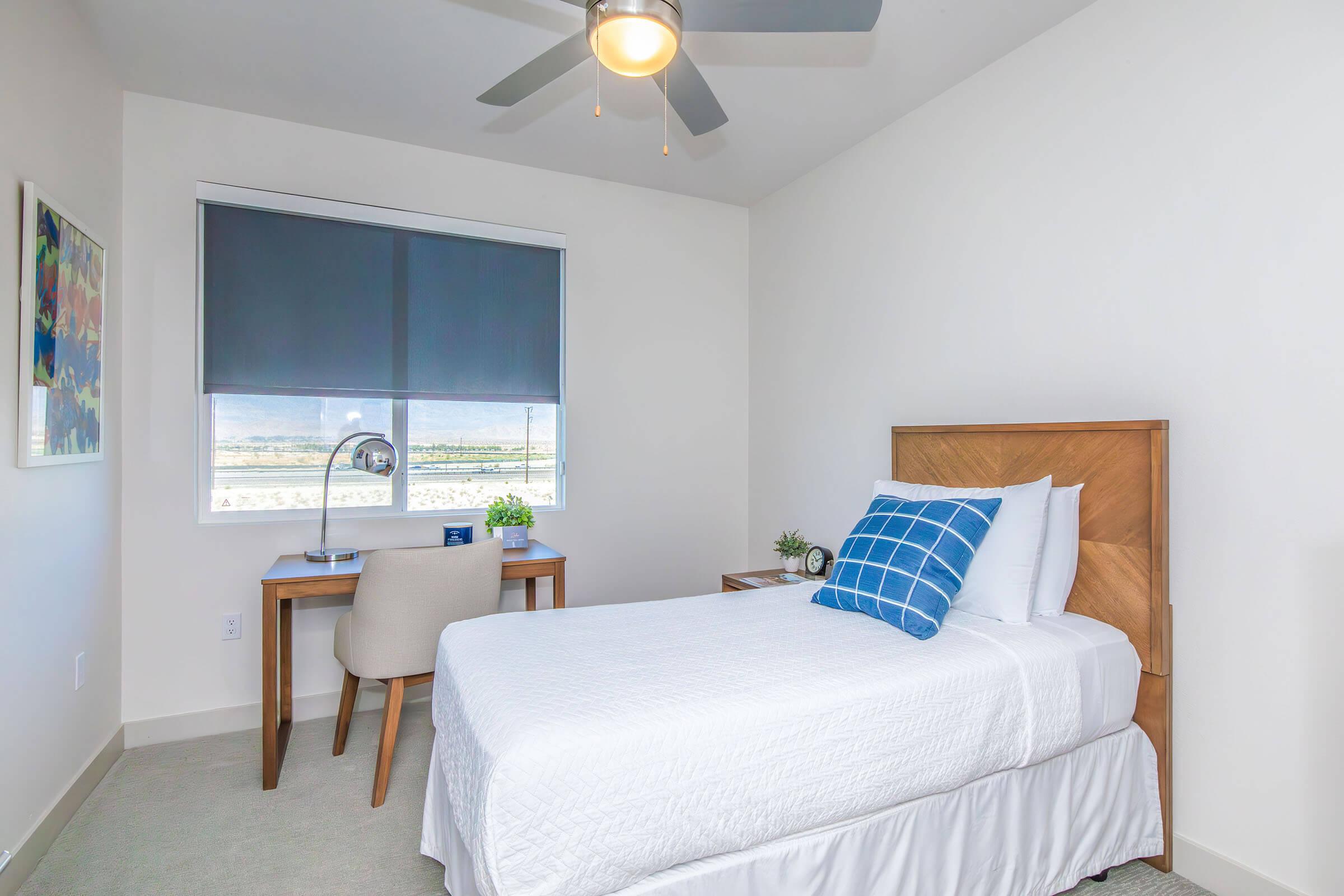 A cozy bedroom featuring a single bed with white bedding, a blue decorative pillow, and a wooden headboard. There's a small desk with a chair, a lamp, and a plant, positioned next to a window with roller shades. The room has a minimalist design and is well-lit, with a view outside.