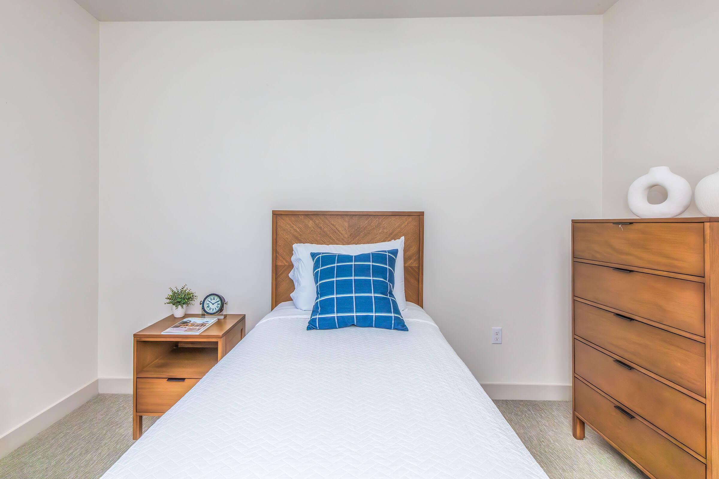 A minimalist bedroom featuring a single bed with a blue checkered pillow, a wooden nightstand with an alarm clock and a small plant, and a tall wooden dresser. The walls are white, and the decor is simple and modern, creating a serene and inviting atmosphere.