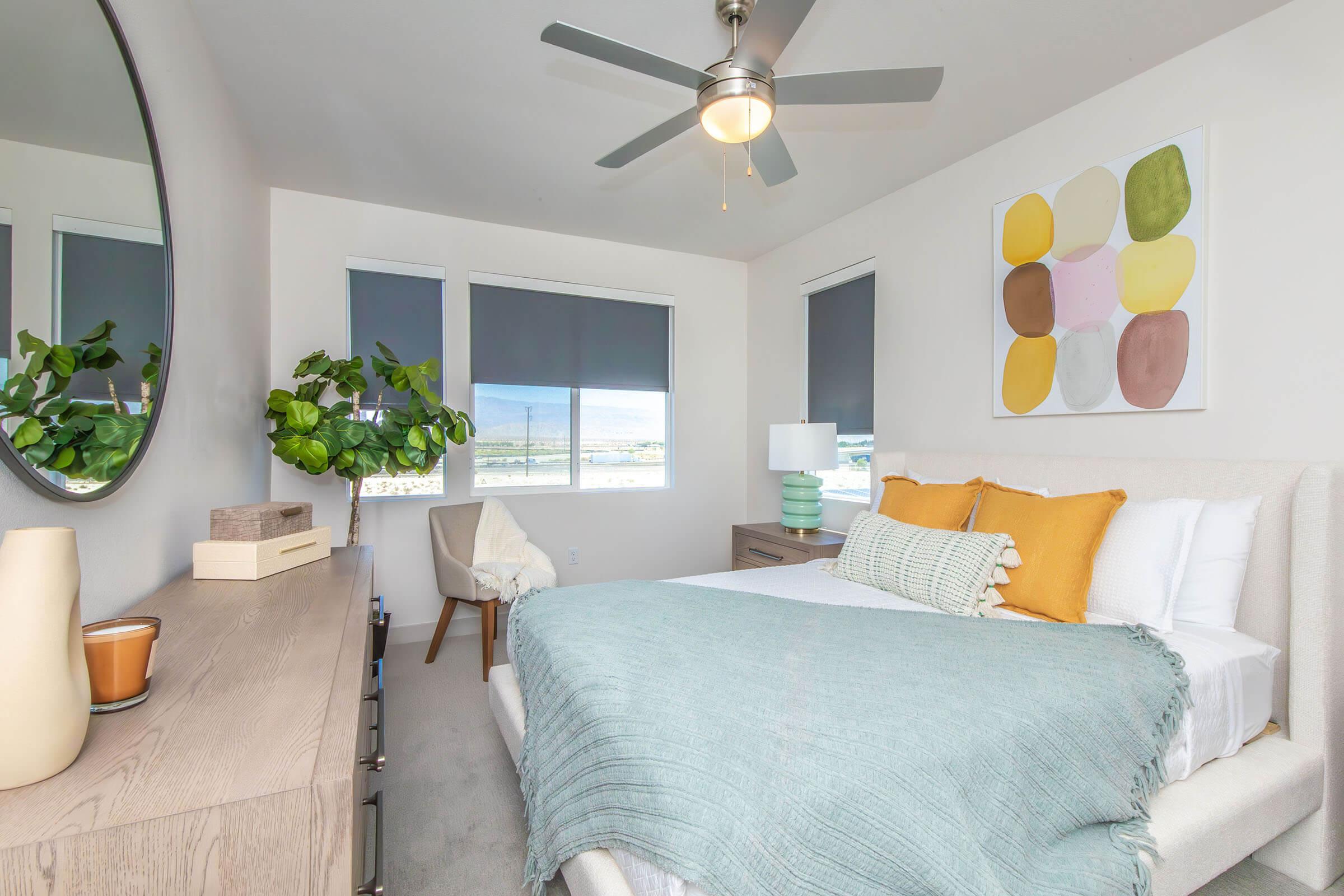 Cozy bedroom featuring a light-colored bed with a teal throw and orange accent pillows. A round mirror hangs above a wooden dresser. There’s a small potted plant, a bedside lamp, and a modern chair nearby. Large windows with blinds offer a view of the outside. A colorful abstract artwork is displayed on the wall.