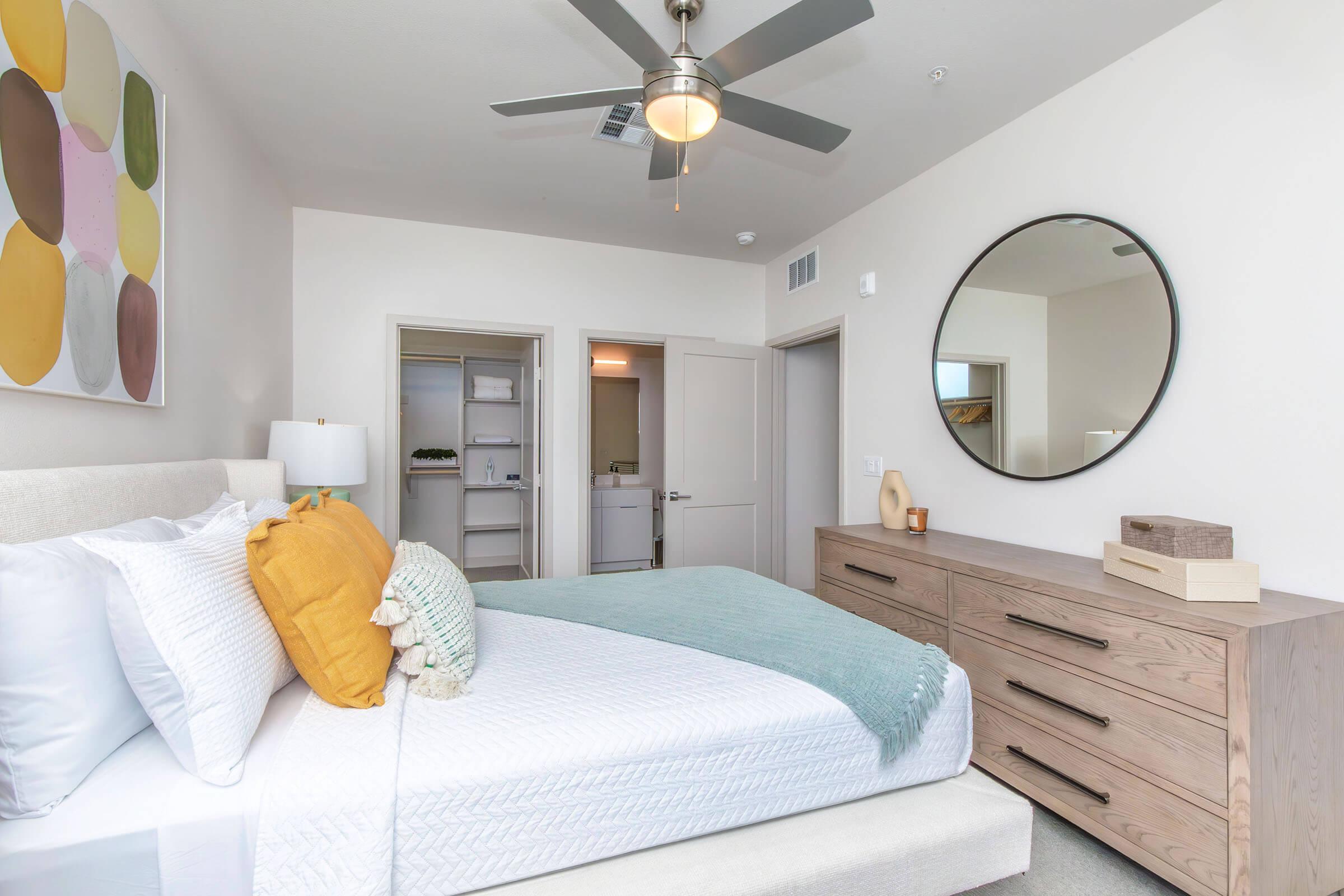 A well-decorated bedroom featuring a comfortable bed with white and yellow bedding, a light blue throw blanket, and a round mirror above a wooden dresser. There's a ceiling fan and abstract wall art with colorful shapes. The room has a neutral color scheme and light-filled spaces, with a door leading to another area.