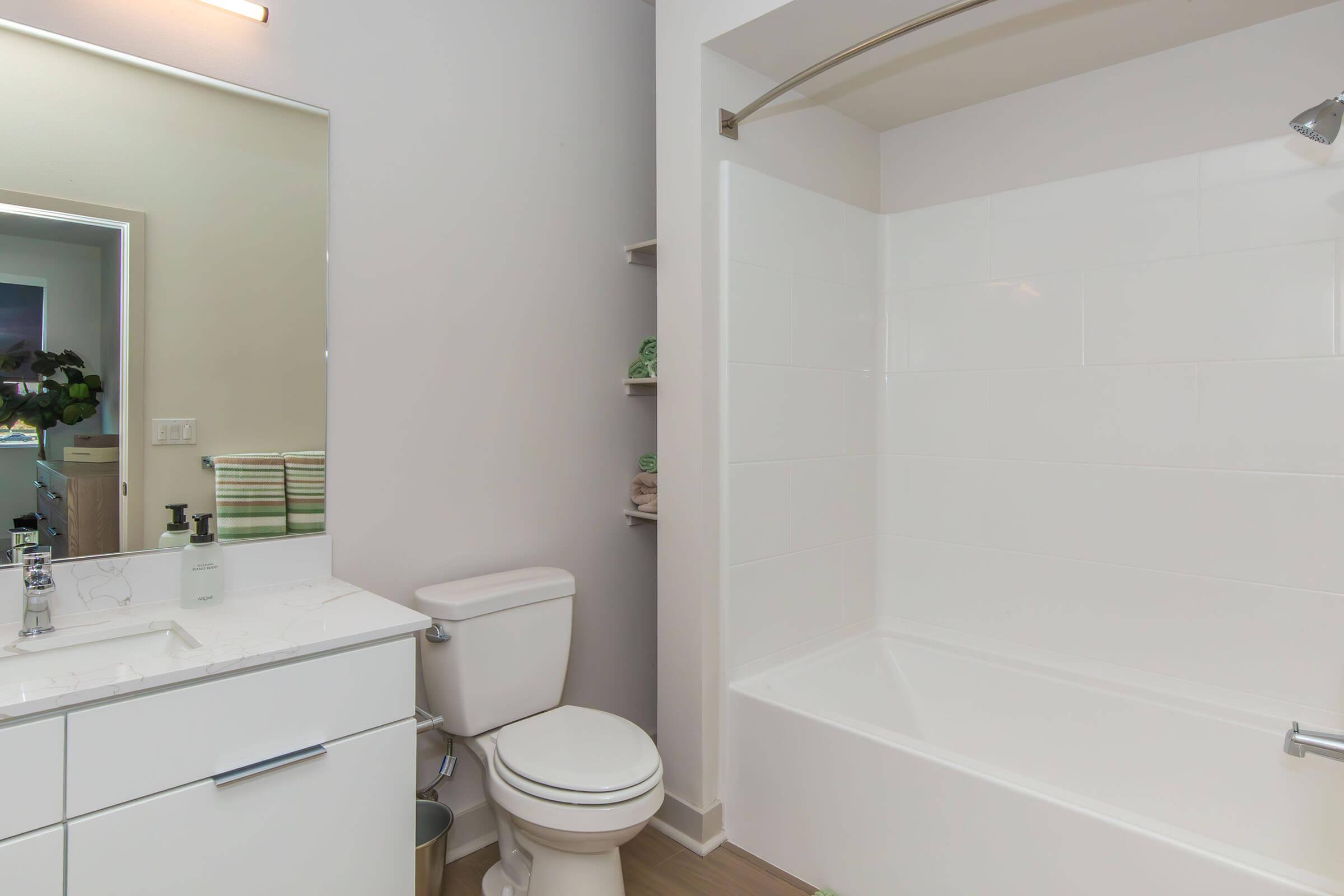 A modern bathroom featuring a white bathtub with a showerhead, a sleek vanity with a mirror, and a toilet. The space has neutral walls and light wood flooring, with neatly stacked towels visible on a shelf in the background.