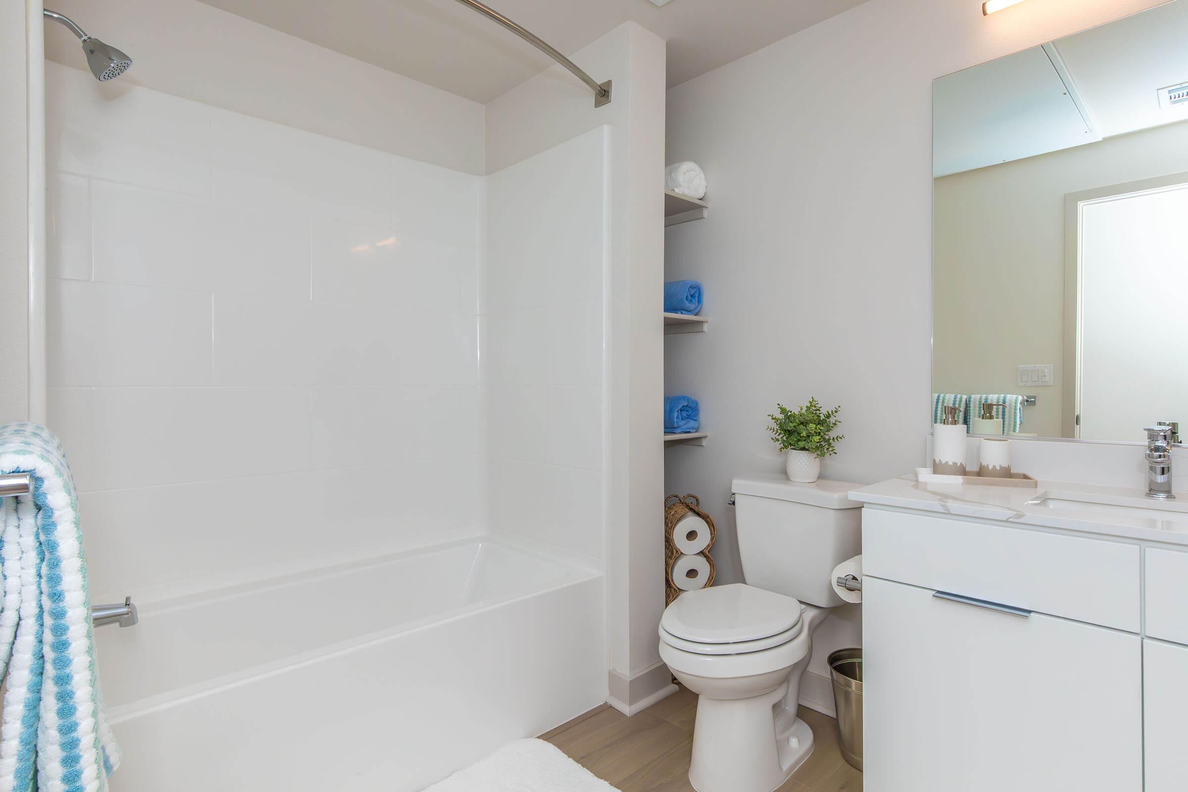 A modern and clean bathroom featuring a white tub and shower combo, a white vanity with a sink, and a toilet. Shelves hold neatly stacked blue towels, and a small plant adds a touch of greenery. Rolled toilet paper is visible, and the overall decor is simple and minimalist.