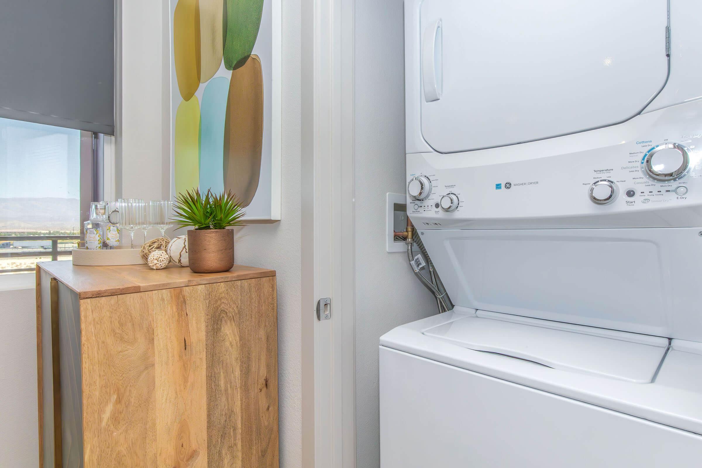A laundry area featuring a stacked washer and dryer next to a wooden cabinet. A small potted plant and decorative items are displayed on top of the cabinet, and there is a colorful wall art piece nearby. Bright natural light enters through a window, illuminating the space.