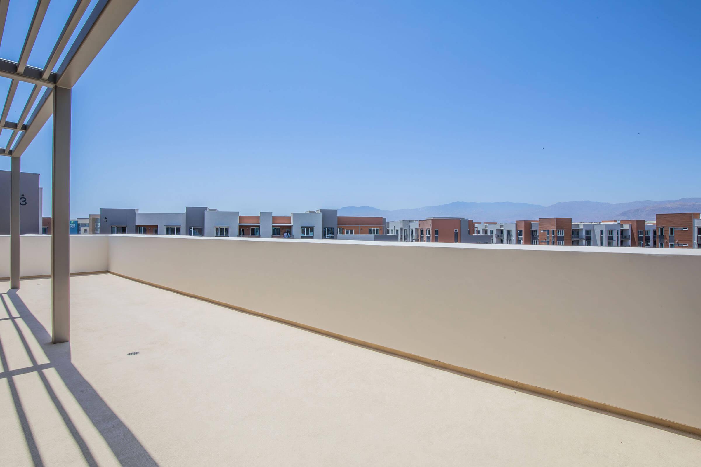 A spacious, empty balcony with a clear view of a blue sky and distant mountains. The foreground features a light-colored railing and the background shows modern buildings with various colors and designs. The scene conveys a sense of openness and tranquility.