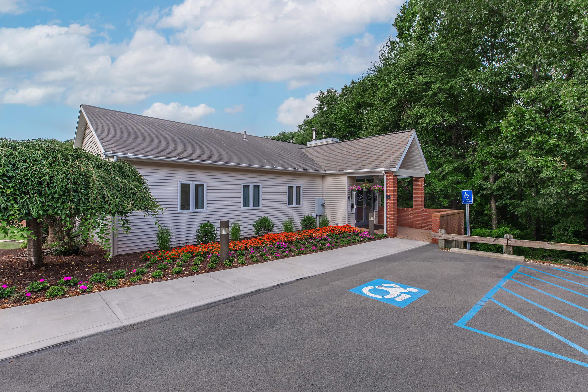 A modern single-story building surrounded by landscaped gardens featuring colorful flowers. A concrete pathway leads to the entrance, which has large windows and a welcoming entrance. Accessible parking spaces are marked in front. Trees line the background, under a partly cloudy sky.