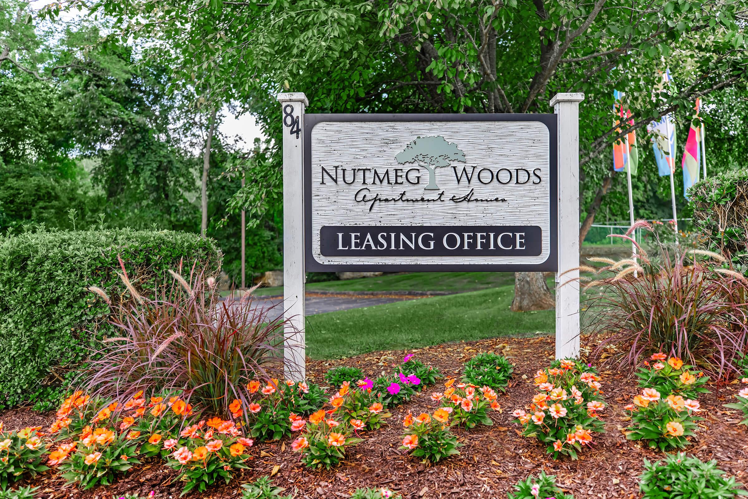 Sign for Nutmeg Woods Apartments Leasing Office, featuring colorful flower beds in front. The sign includes the name of the apartments and indicates the leasing office's location. Lush greenery surrounds the area, creating a welcoming atmosphere.