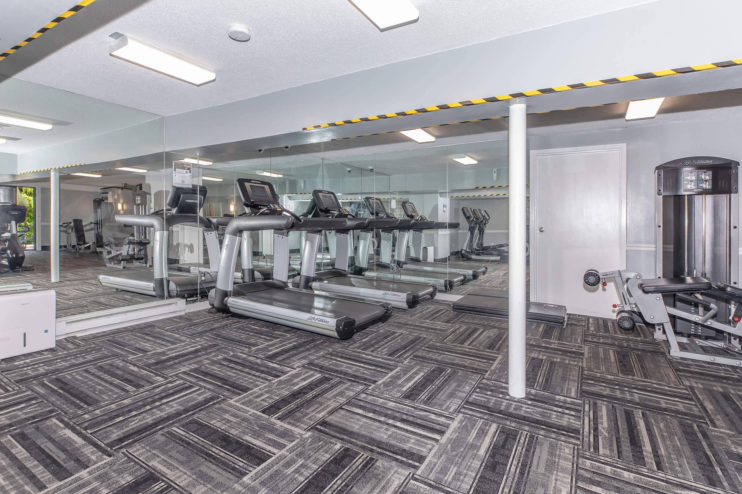 A modern gym interior featuring several treadmills lined up against a mirrored wall. The space has a gray and black patterned carpet and includes various fitness equipment. Bright overhead lighting enhances the open feel of the room, making it inviting for workouts. There are caution tape markings along the ceiling.