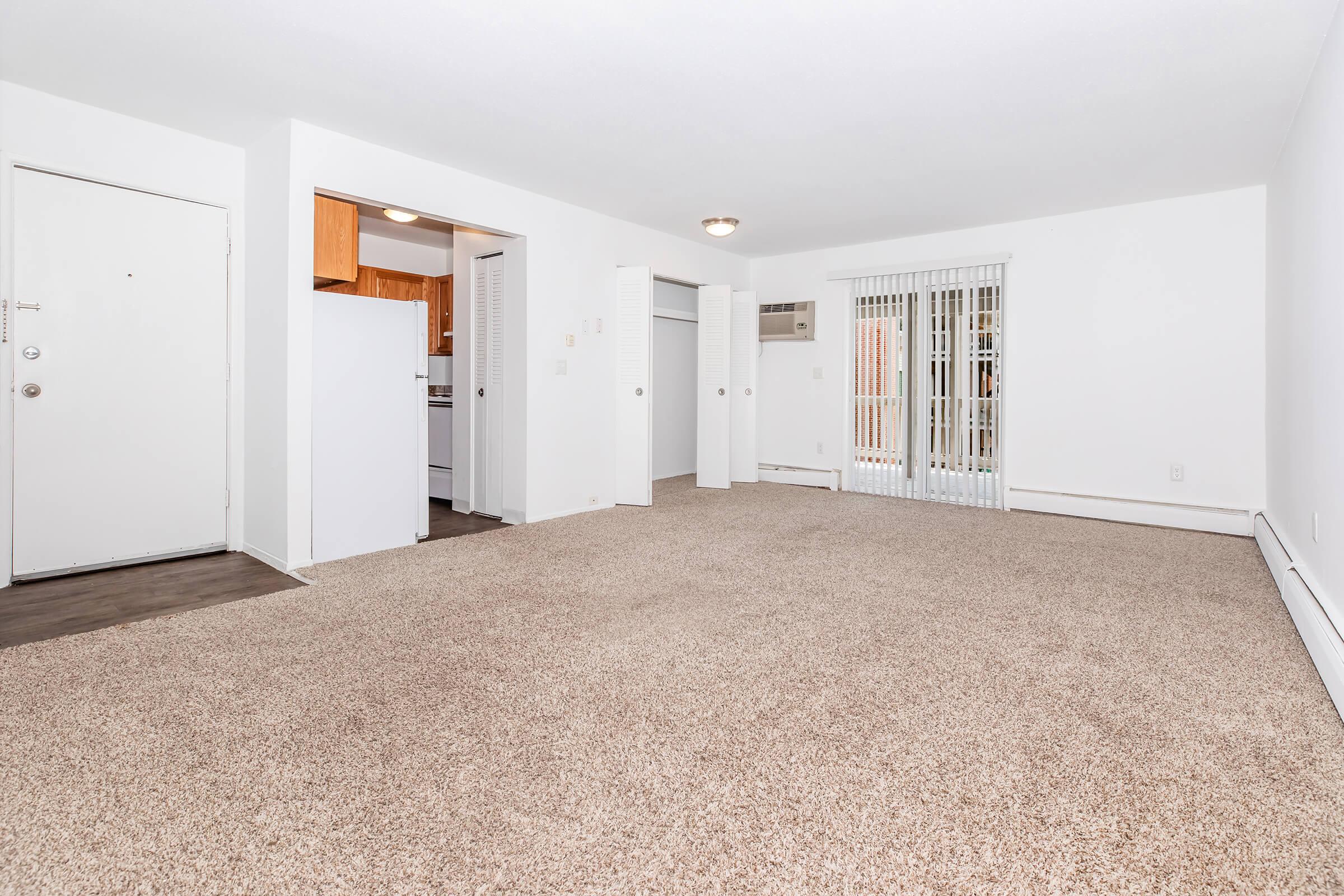 A spacious, well-lit living area with light beige carpet, featuring a door leading outside and an open kitchen area. There is a small hallway leading to additional rooms and an air conditioning unit on the wall. The walls are painted white, creating a clean and modern aesthetic.