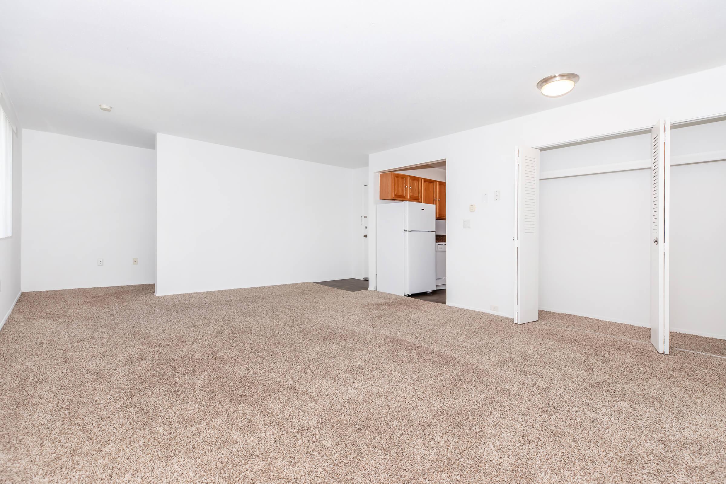A spacious, empty room with light-colored walls and carpet flooring. On one side, there are two sliding closet doors and an opening leading to a kitchen area with wooden cabinets and a white refrigerator visible in the background. Natural light comes in through a window on the left.