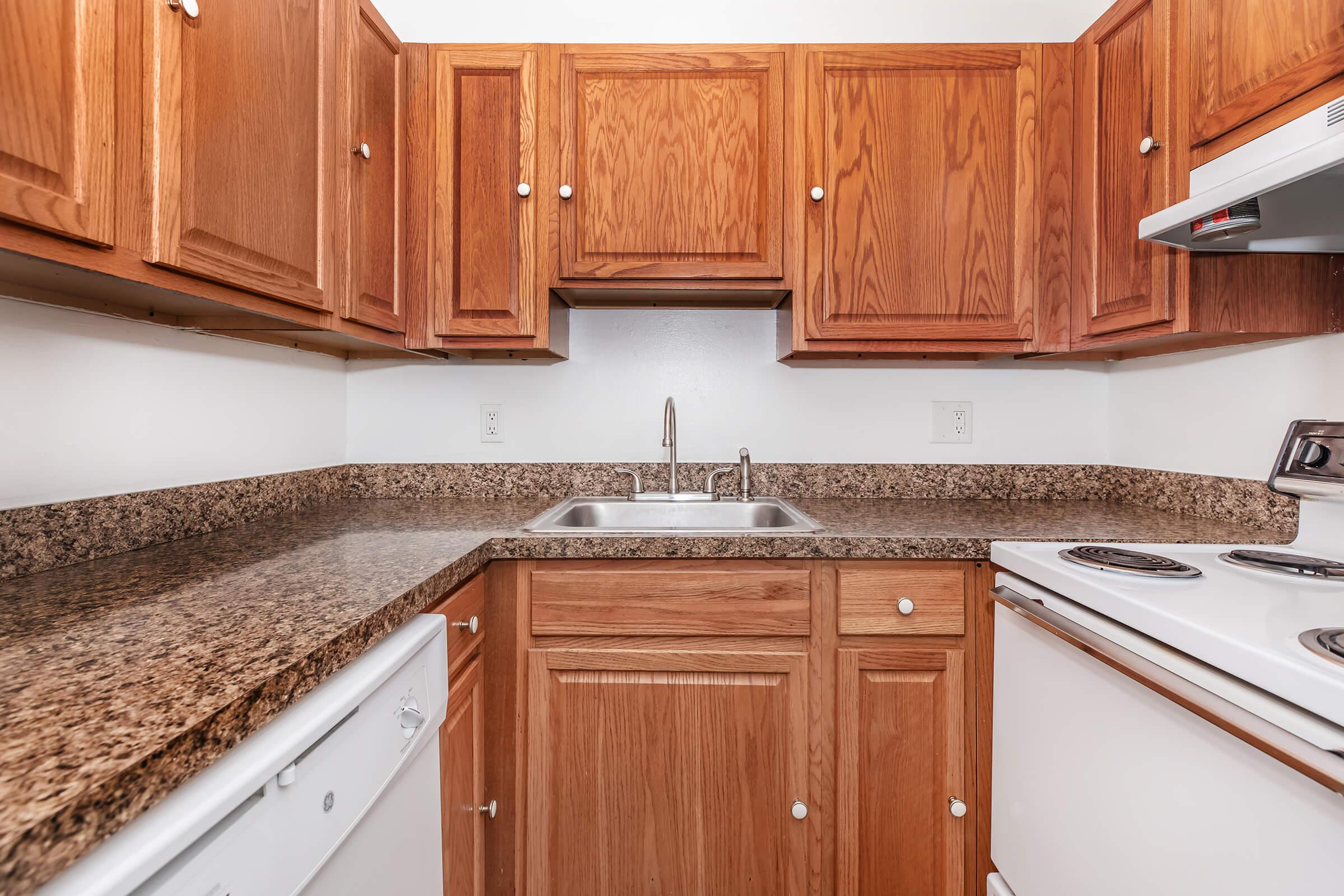 A small kitchen featuring wooden cabinets above a granite countertop. There is a stainless steel sink with a faucet, a white dishwasher, and a white stove with an oven. The walls are painted a light color, creating a bright and clean atmosphere.