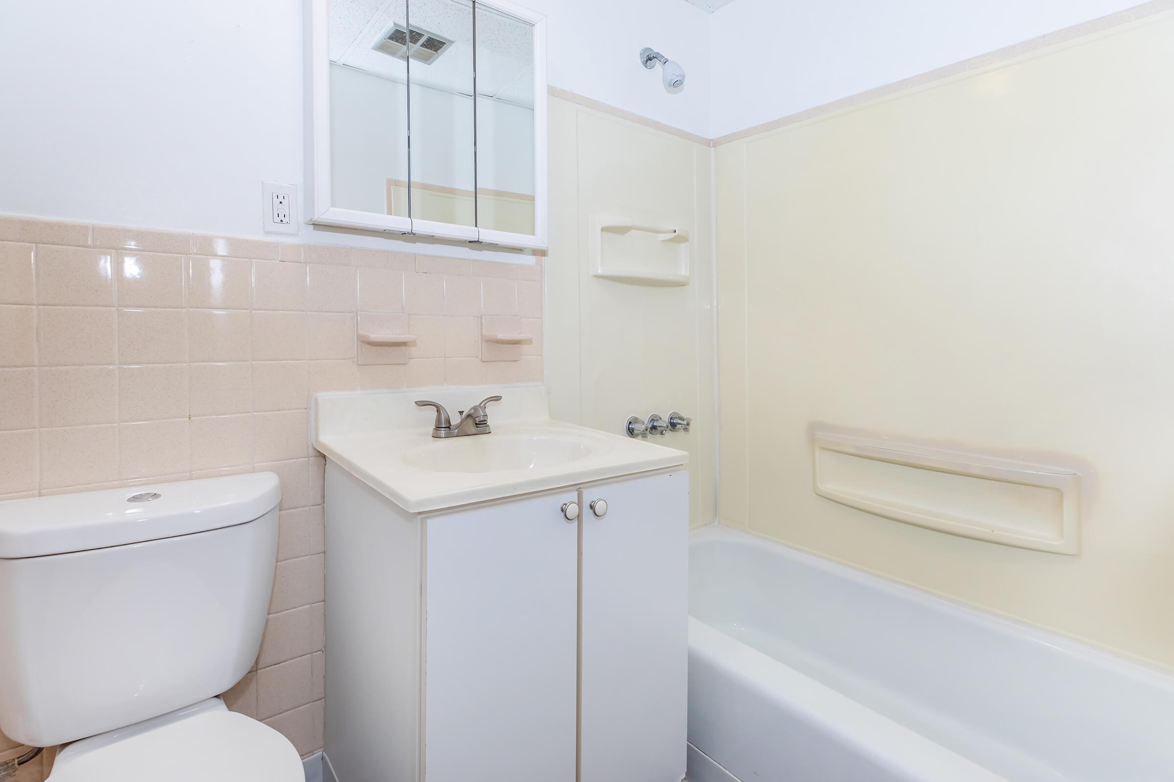 A clean, neutral-colored bathroom featuring a white toilet, a small sink with a mirror above it, and a bathtub. The walls are mostly light yellow with beige tiles. The space appears well-lit and tidy, suitable for everyday use.