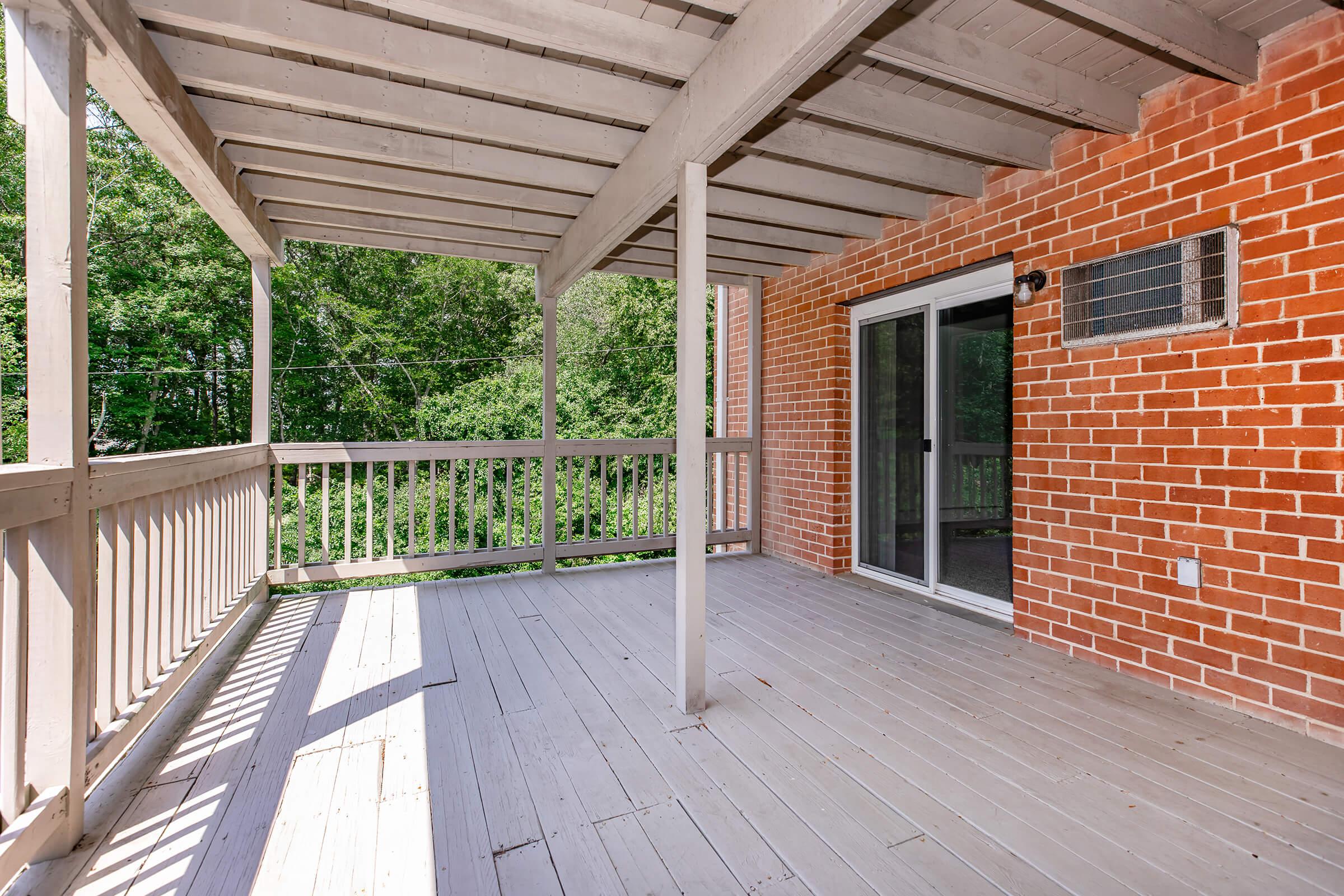 A spacious outdoor balcony with wooden flooring and a covered roof, featuring a brick wall and a glass door leading indoors. Lush greenery is visible in the background, creating a serene atmosphere. The space is well-lit by natural sunlight.