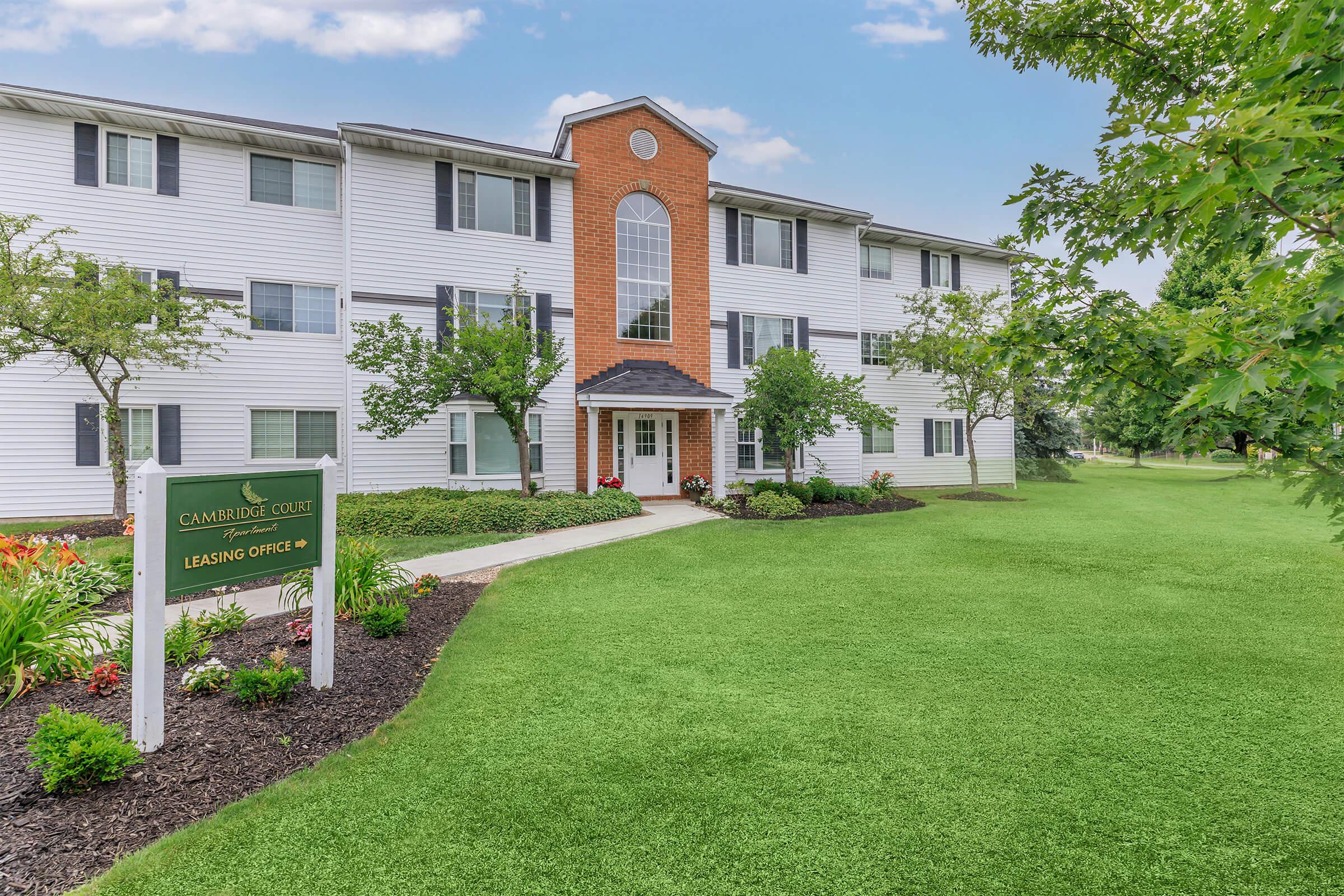 Exterior view of a residential apartment complex with a well-maintained lawn and landscaping. The building features multiple stories with a brick and white siding façade. A sign indicating "Cambridge Court Leasing Office" is prominently displayed near the entrance. Trees and shrubs enhance the green space around the entrance.