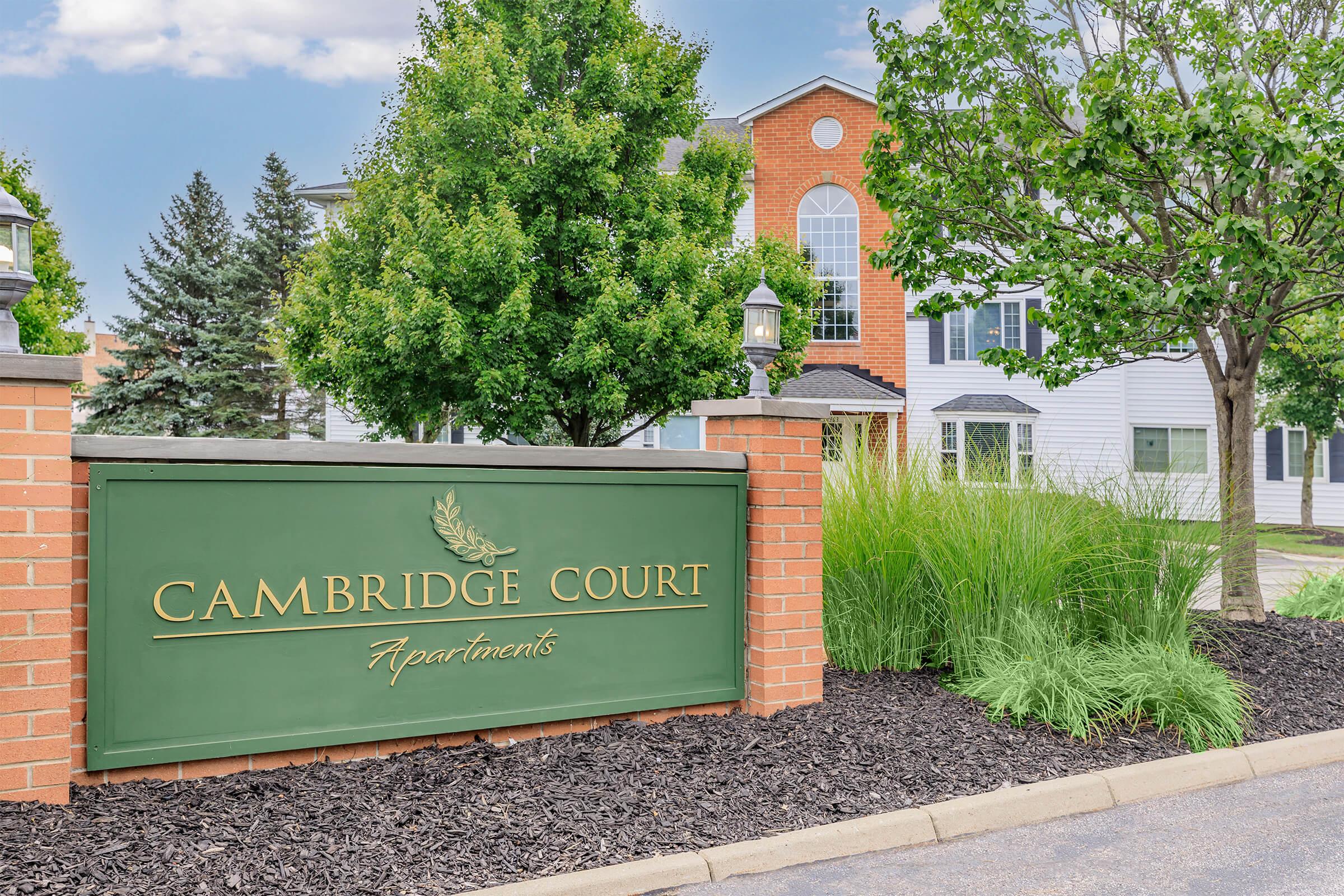 Sign for Cambridge Court Apartments, featuring a decorative green background with the name in gold lettering. Surrounding the sign are well-maintained shrubs and trees, with a modern apartment building visible in the background under a clear blue sky.