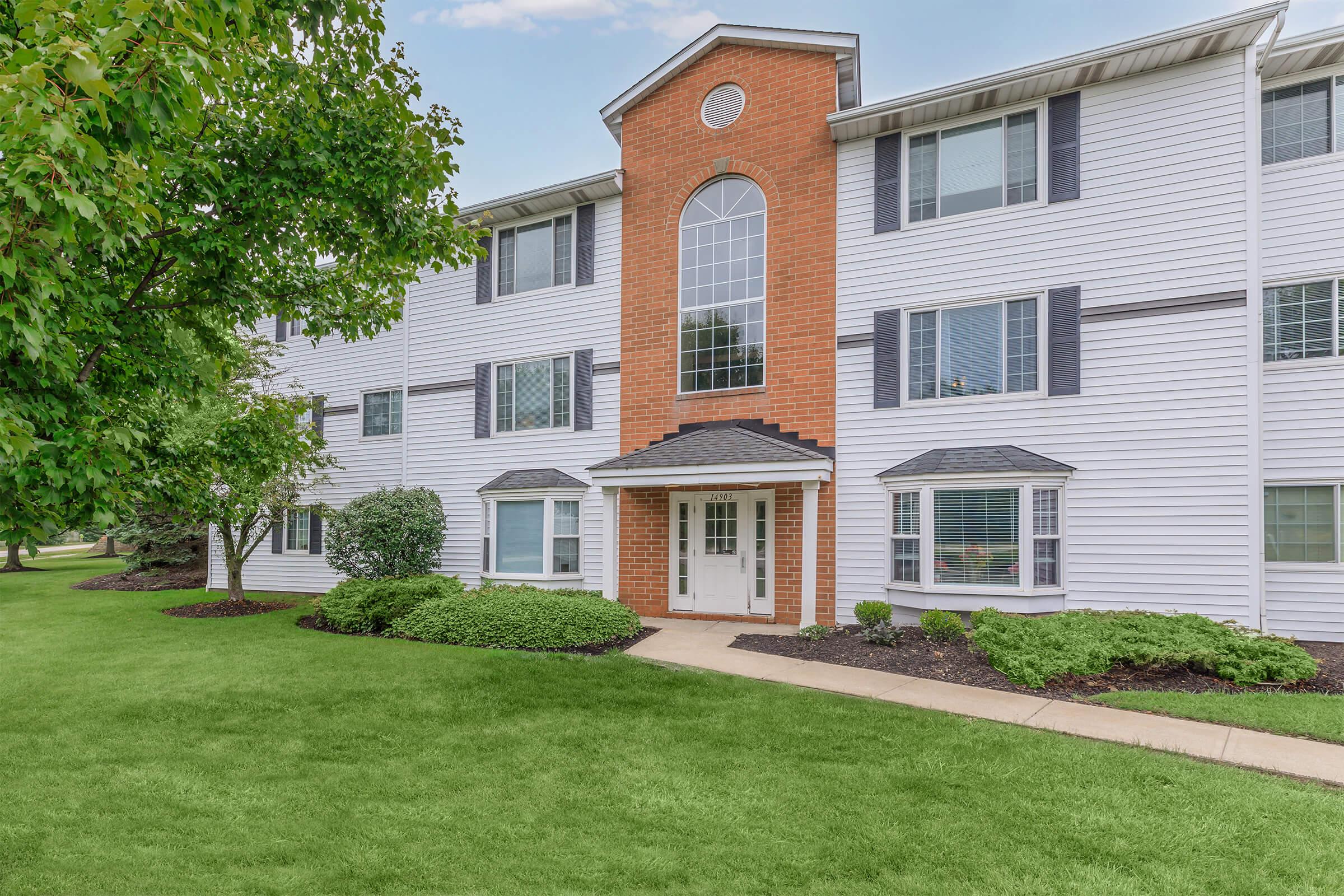 A modern apartment building with a brick and white exterior. It features large windows, a prominent entrance with double doors, and landscaped greenery including shrubs and a lawn. The facade is complemented by a symmetrical design with an arched window above the entrance.