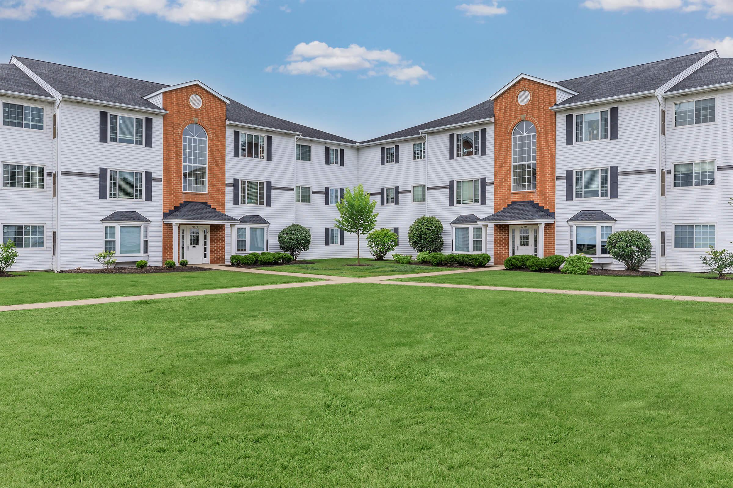 A view of a two-story apartment building with white siding and red brick accents, surrounded by manicured green lawns and small bushes. The building features several large windows and a symmetrical design, with a pathway leading through the grass, creating an inviting atmosphere.