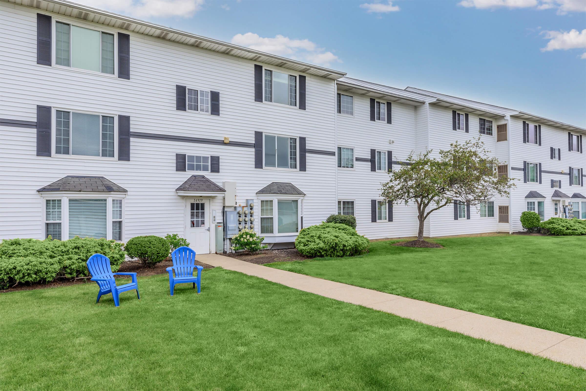 A well-maintained apartment complex featuring a grassy lawn. Two blue adirondack chairs are positioned on the lawn in front of the building, which has multiple windows and a white exterior. The sky is partly cloudy, adding to the overall inviting atmosphere of the scene.