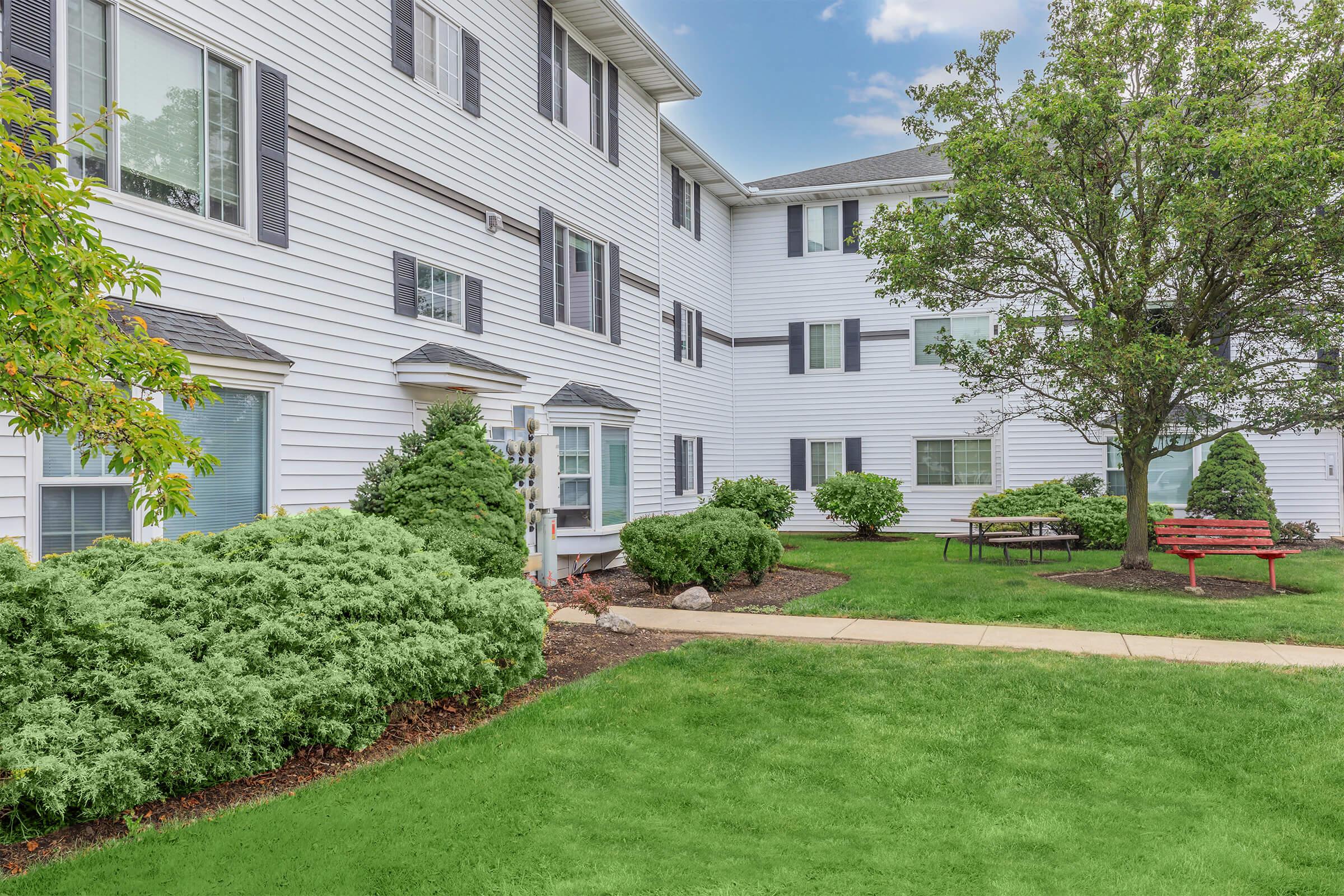 A landscaped area featuring a multi-story residential building with white siding. Neatly trimmed shrubs and bushes are in the foreground, along with a picnic table and red benches on a green lawn. The sky is clear with a few clouds, creating a welcoming atmosphere.