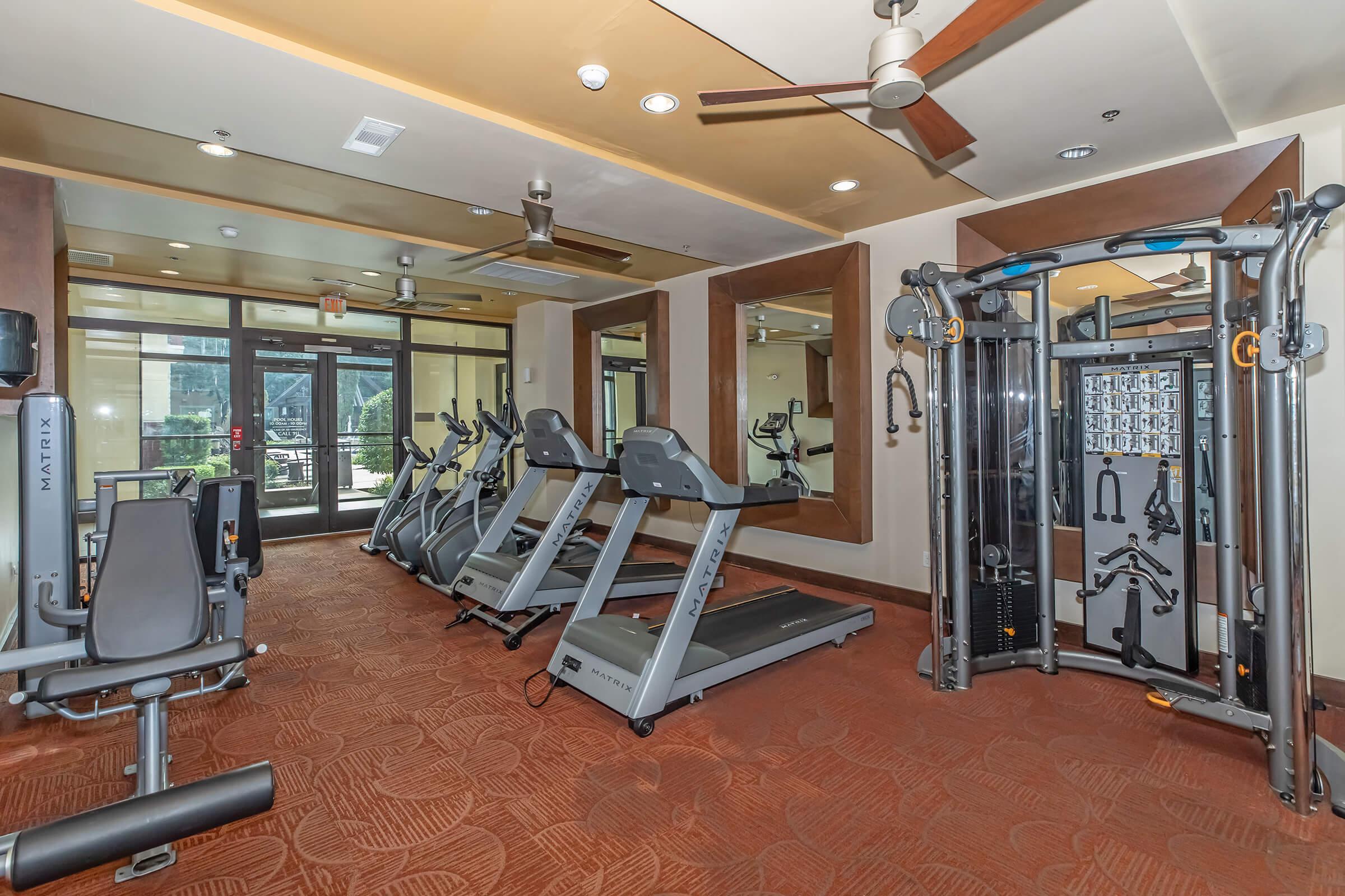 A well-lit fitness room featuring treadmills, elliptical machines, and a multi-station workout machine. Large mirrors reflect the equipment, and there are windows providing natural light. The room has a modern design with a warm color scheme and ceiling fans.