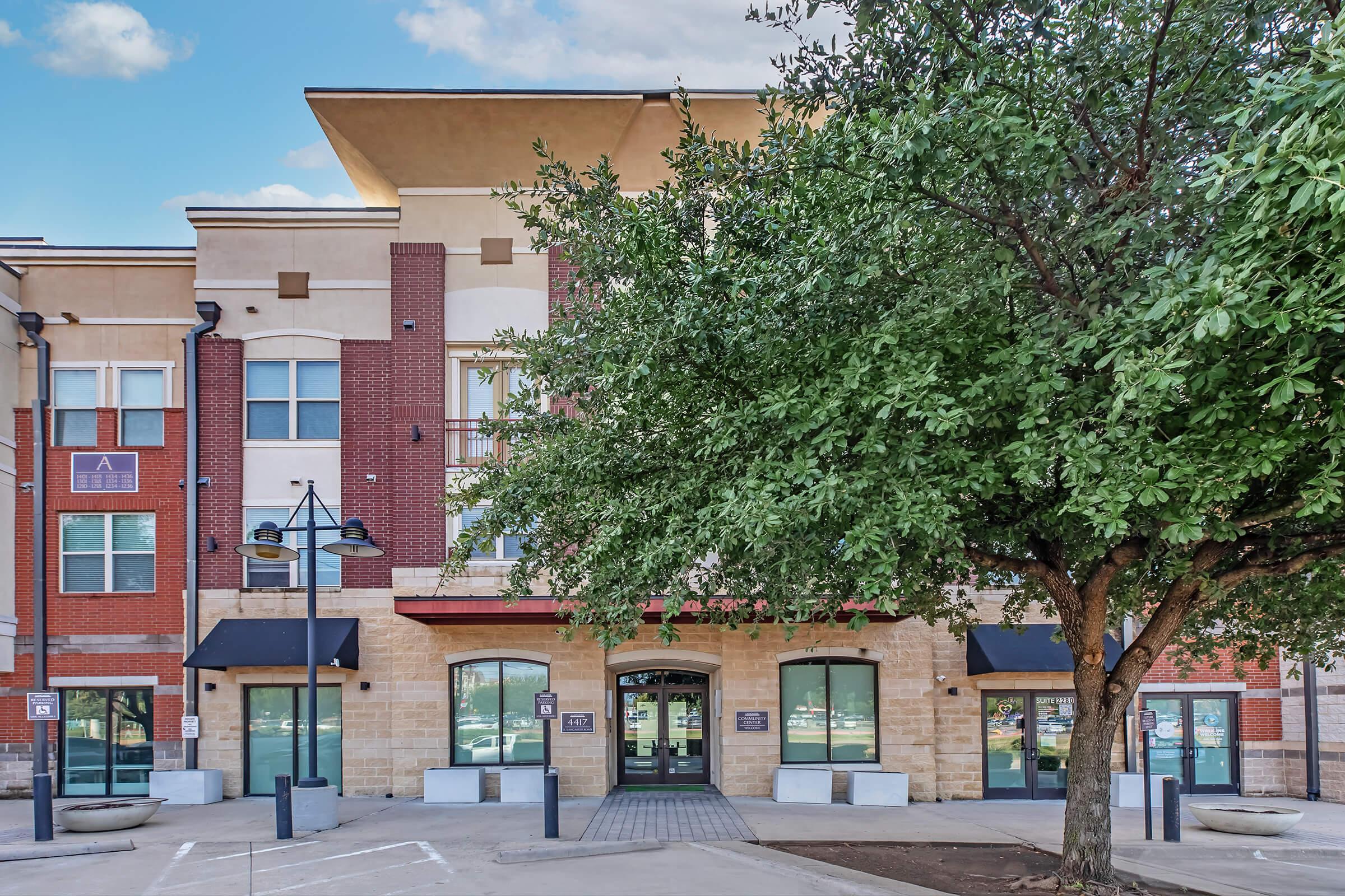 A modern multi-story building with a blend of brick and tan exterior. The entrance features large glass doors and awnings. Surrounding the building are green trees and a parking area. The sky is blue with scattered clouds, creating a bright and welcoming atmosphere.