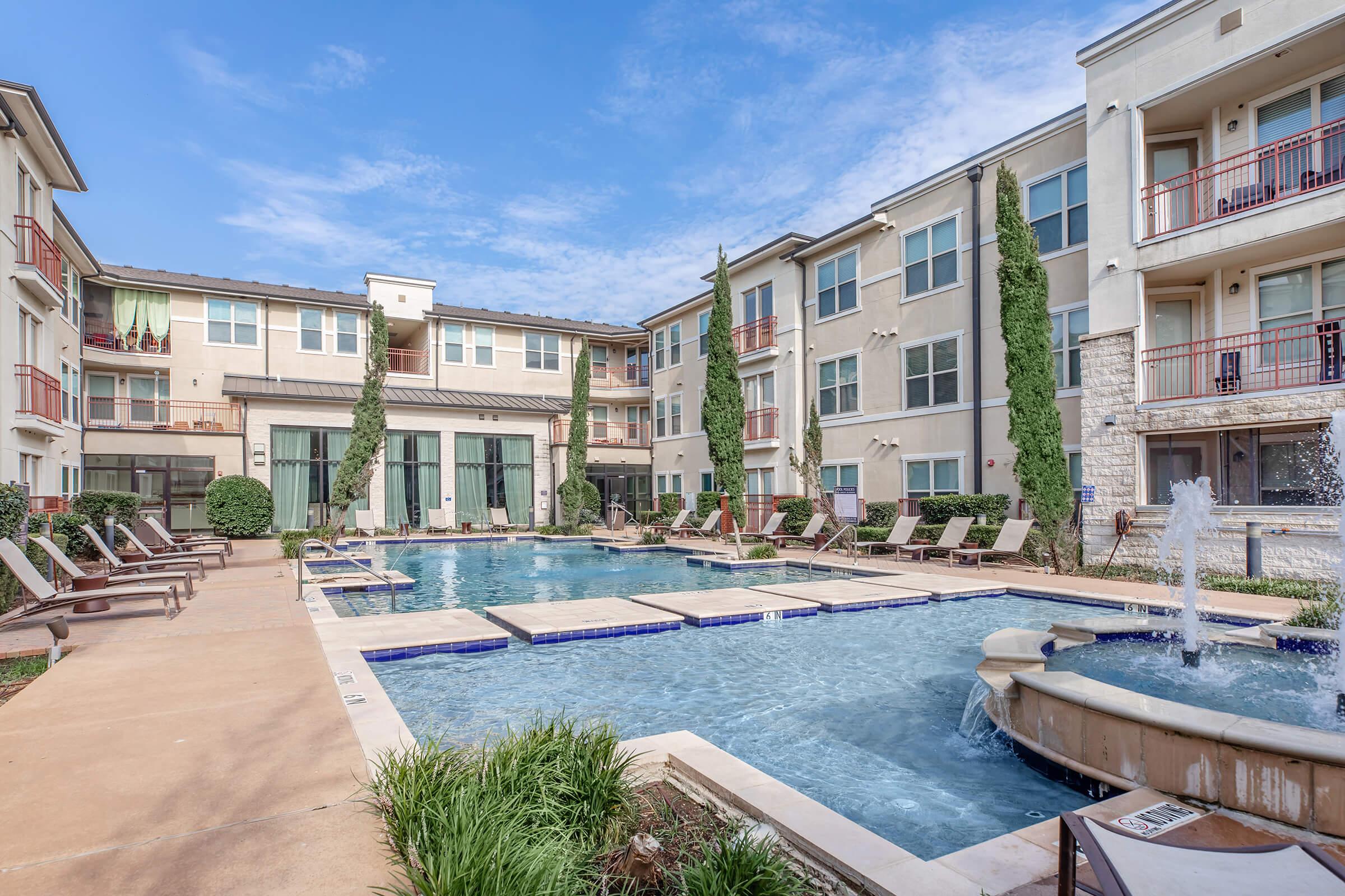 A bright outdoor pool area surrounded by an apartment complex. The pool features lounge chairs, a fountain, and landscaped greenery. Two buildings in the background have balconies and large windows, and the sky is clear with a few clouds.