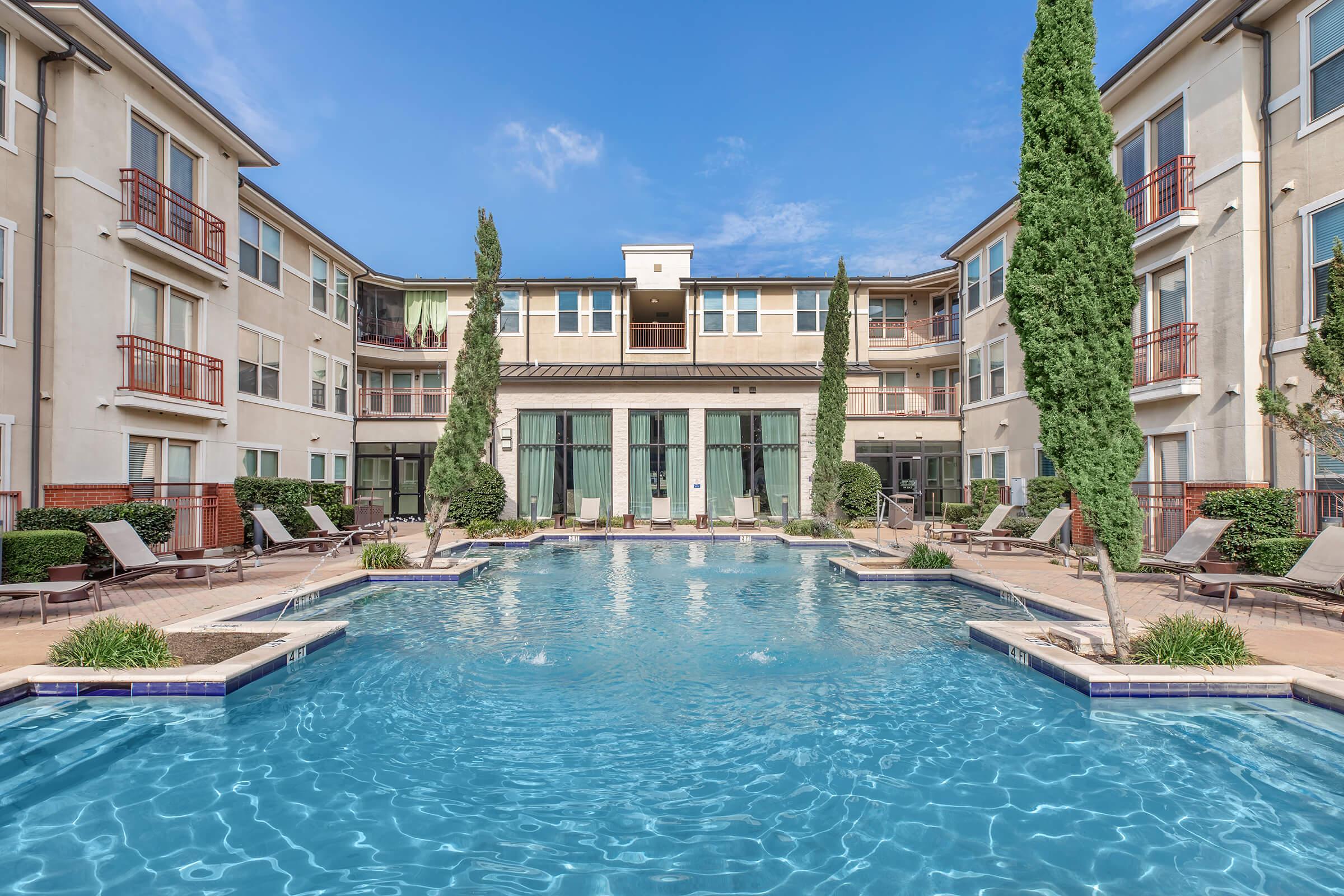 A serene swimming pool surrounded by lounge chairs and tall cypress trees, with a modern apartment building in the background. The clear blue sky reflects on the water, creating a peaceful atmosphere ideal for relaxation and leisure.