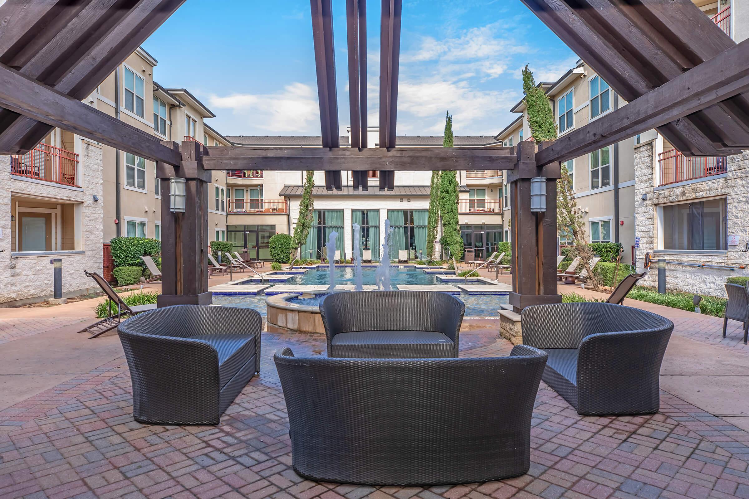 A cozy outdoor seating area with four black wicker chairs arranged around a small circular table, overlooking a serene courtyard with a fountain and well-maintained greenery. The surrounding buildings feature modern architecture and large windows, creating a welcoming atmosphere.