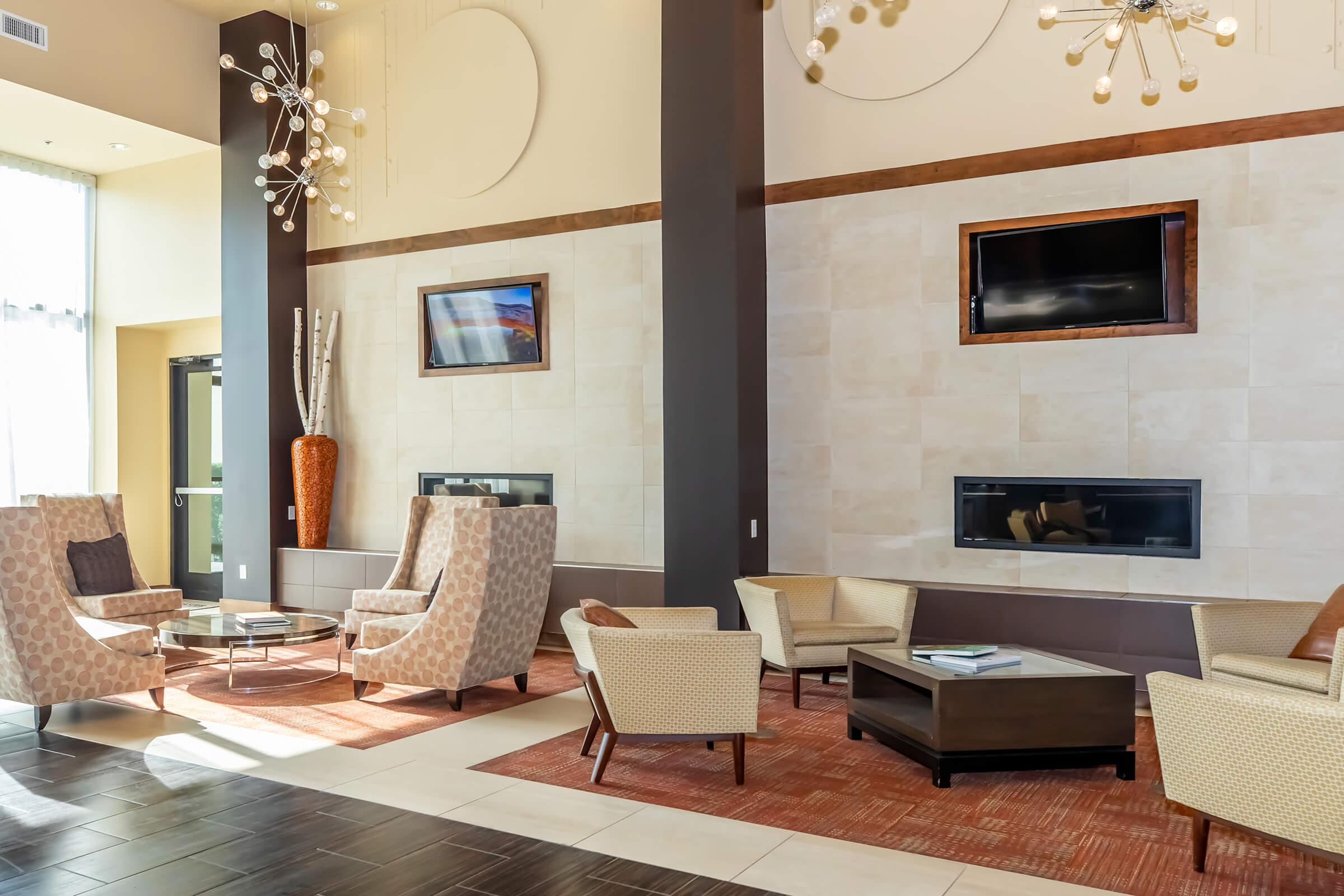 Modern lounge area featuring stylish chairs and a coffee table, with a large window allowing natural light. Elegant decorative elements include a floor vase and contemporary light fixtures. Two wall-mounted TVs are visible, enhancing the inviting atmosphere.